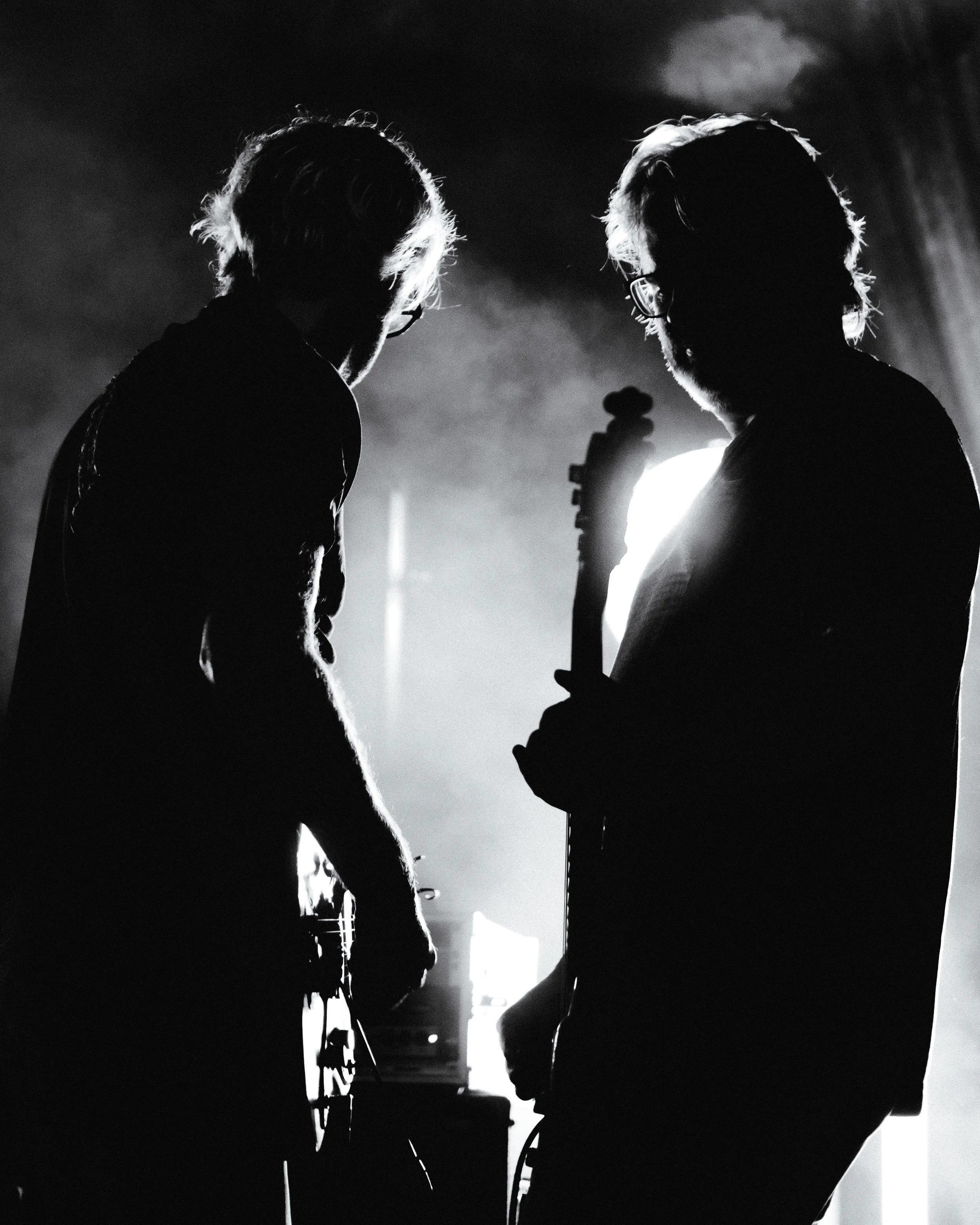 Two musicians with guitars performing on stage in black and white, backlit to create silhouettes.