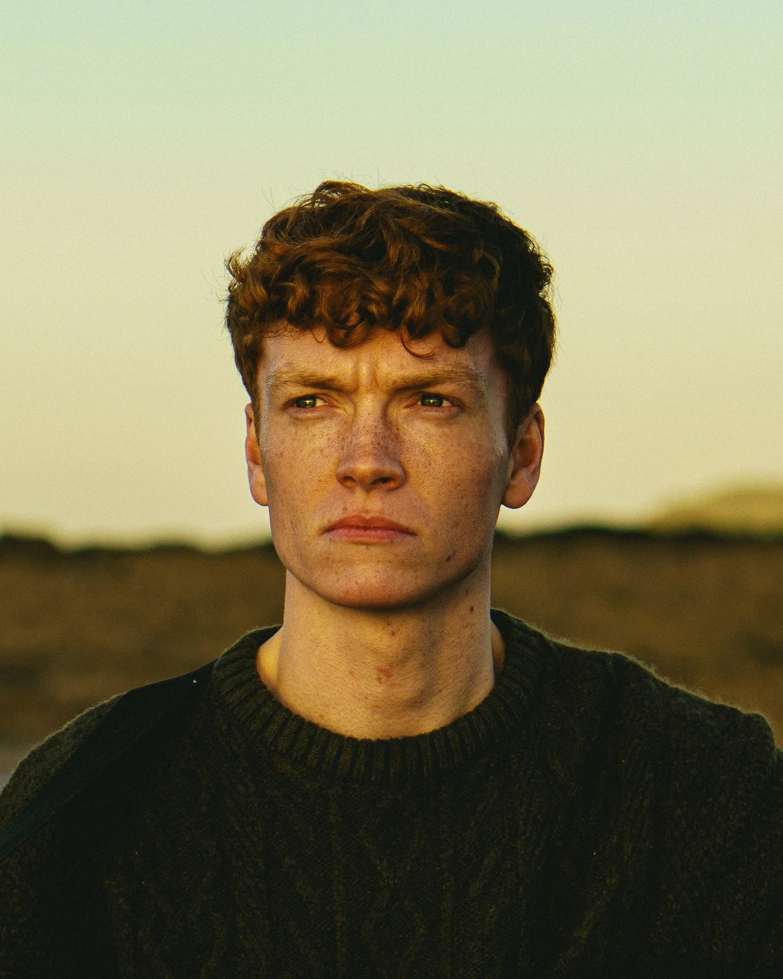 A young man with curly brown hair and fair skin standing outdoors during golden hour, wearing a dark sweater, looking off to the side with a serious expression.