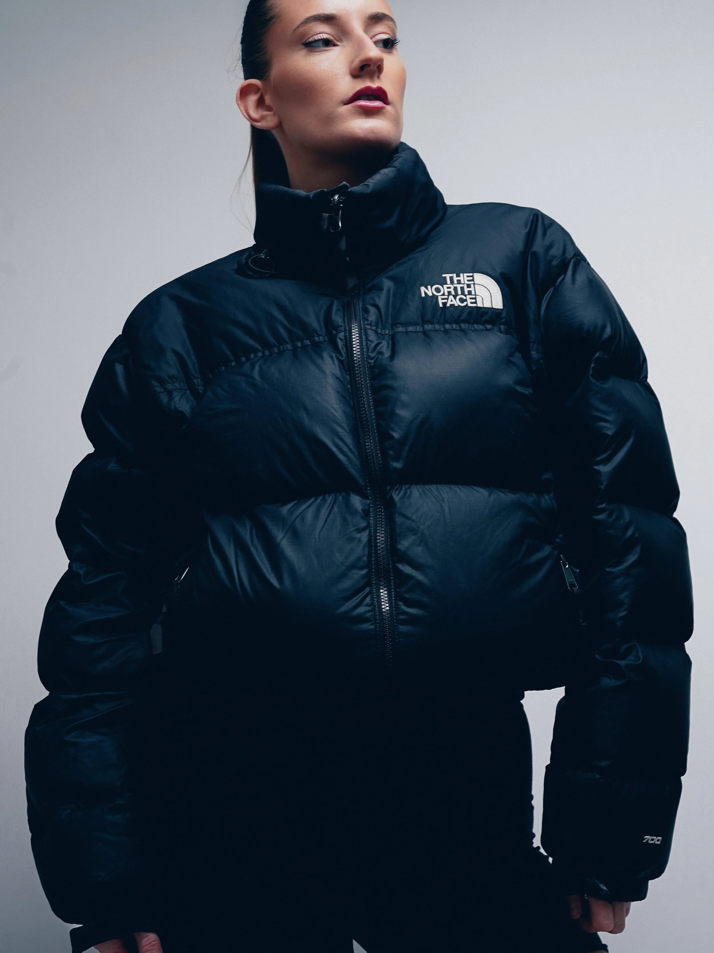 Woman wearing a black North Face puffer jacket with a high collar, standing against a plain background.