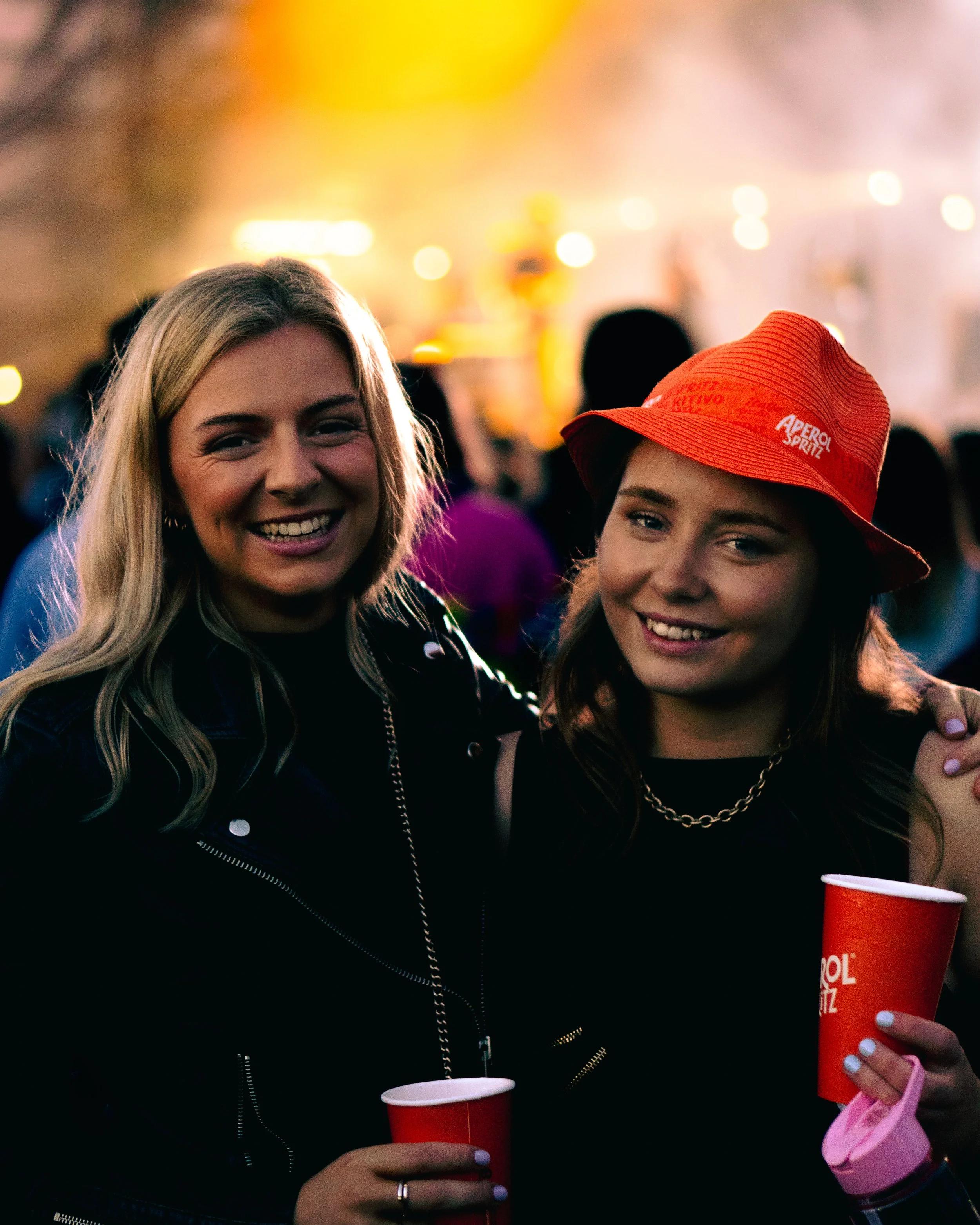 Two women smiling at a social gathering, holding red cups, with blurred lights and other people in the background.
