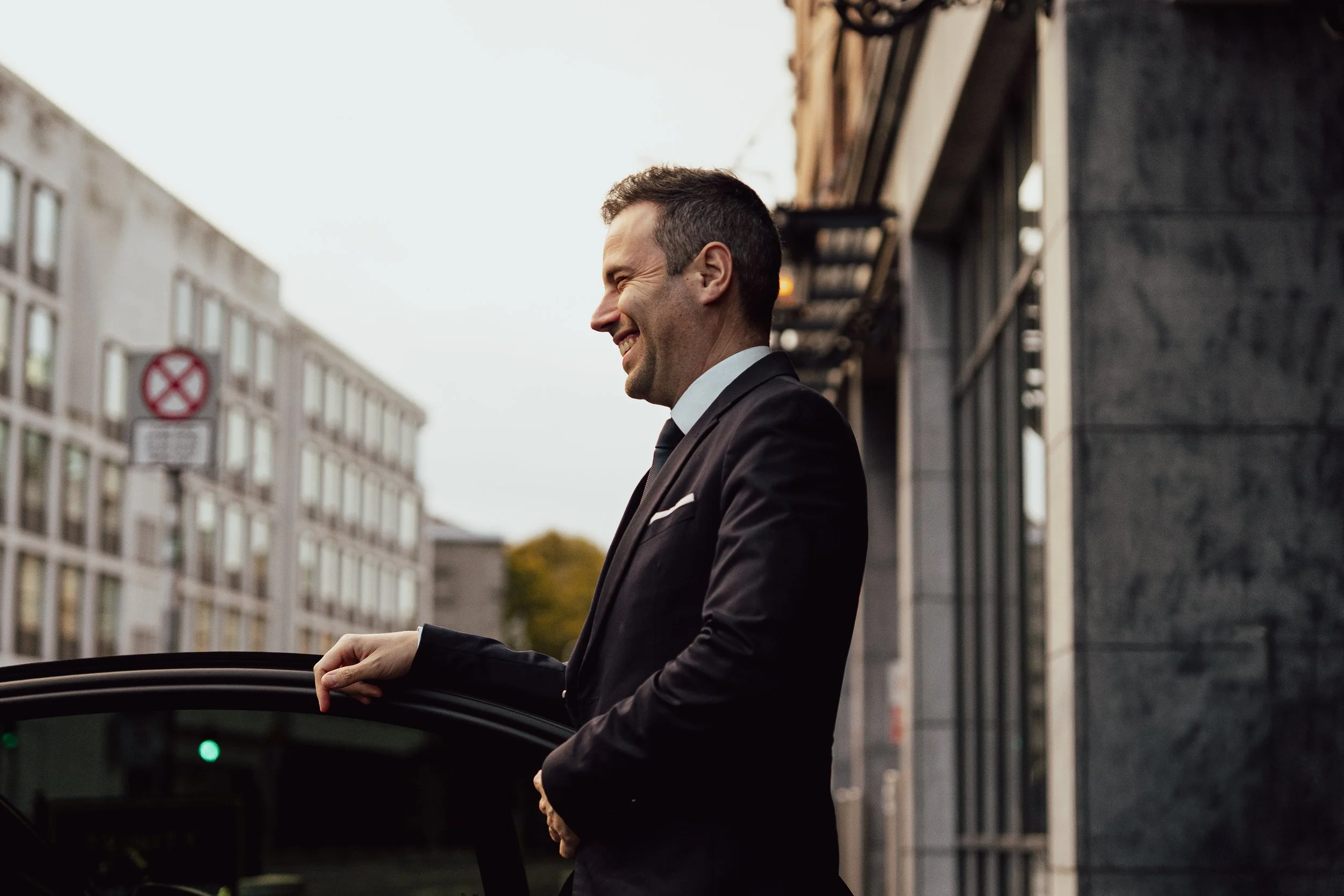 A smiling man in a black suit and tie standing outside near a building, with his hand on the open door of a car.