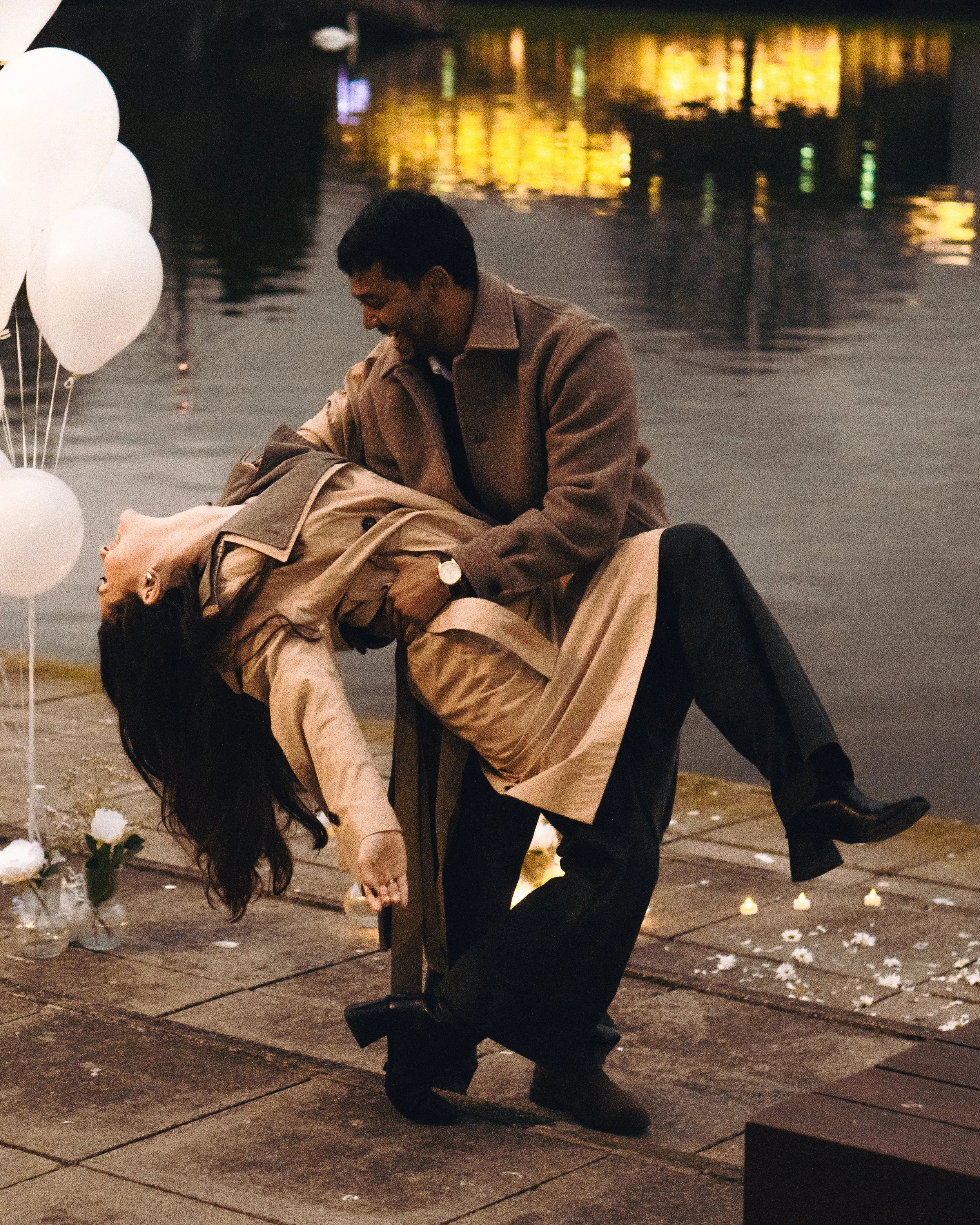 A man holding a woman on a dock decorated with white balloons and flowers, with candles on the ground, near a body of water at sunset.