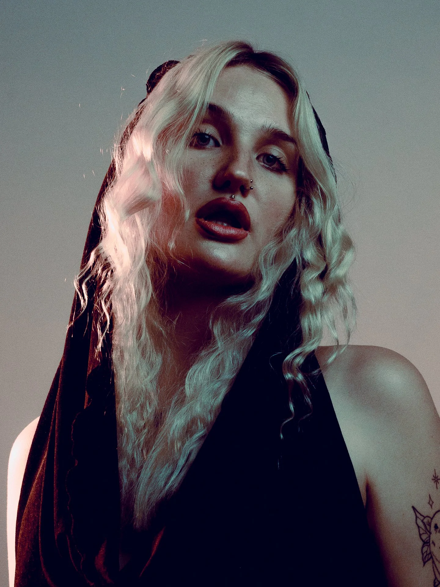 A woman with long curly blonde and black hair, wearing dark lipstick, a septum piercing, and a black top, with a tattoo on her left shoulder, looking into the camera with a slightly open mouth.