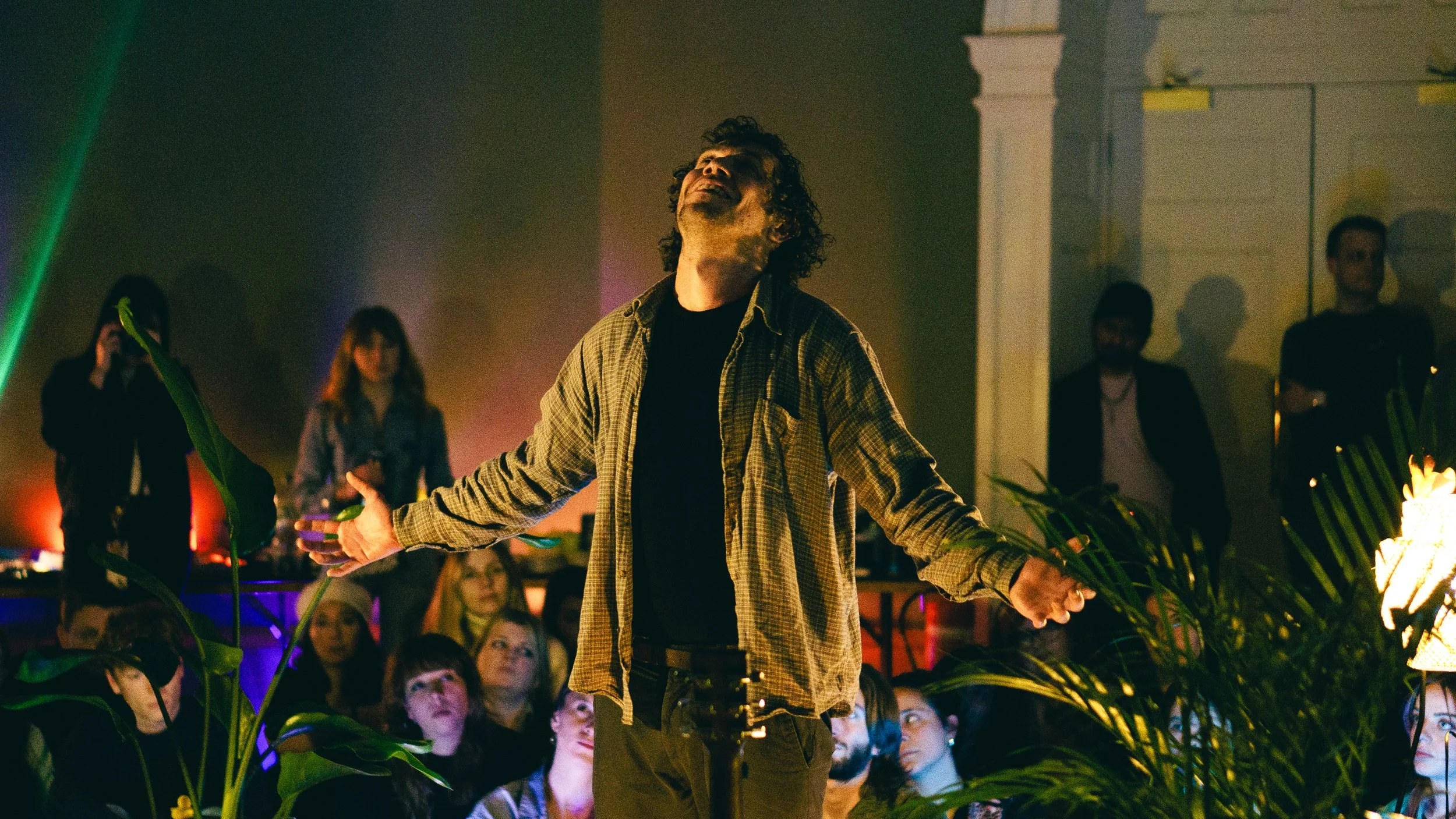 Man performing with arms outstretched in front of an audience at a dimly lit indoor event with plants and colorful lights.