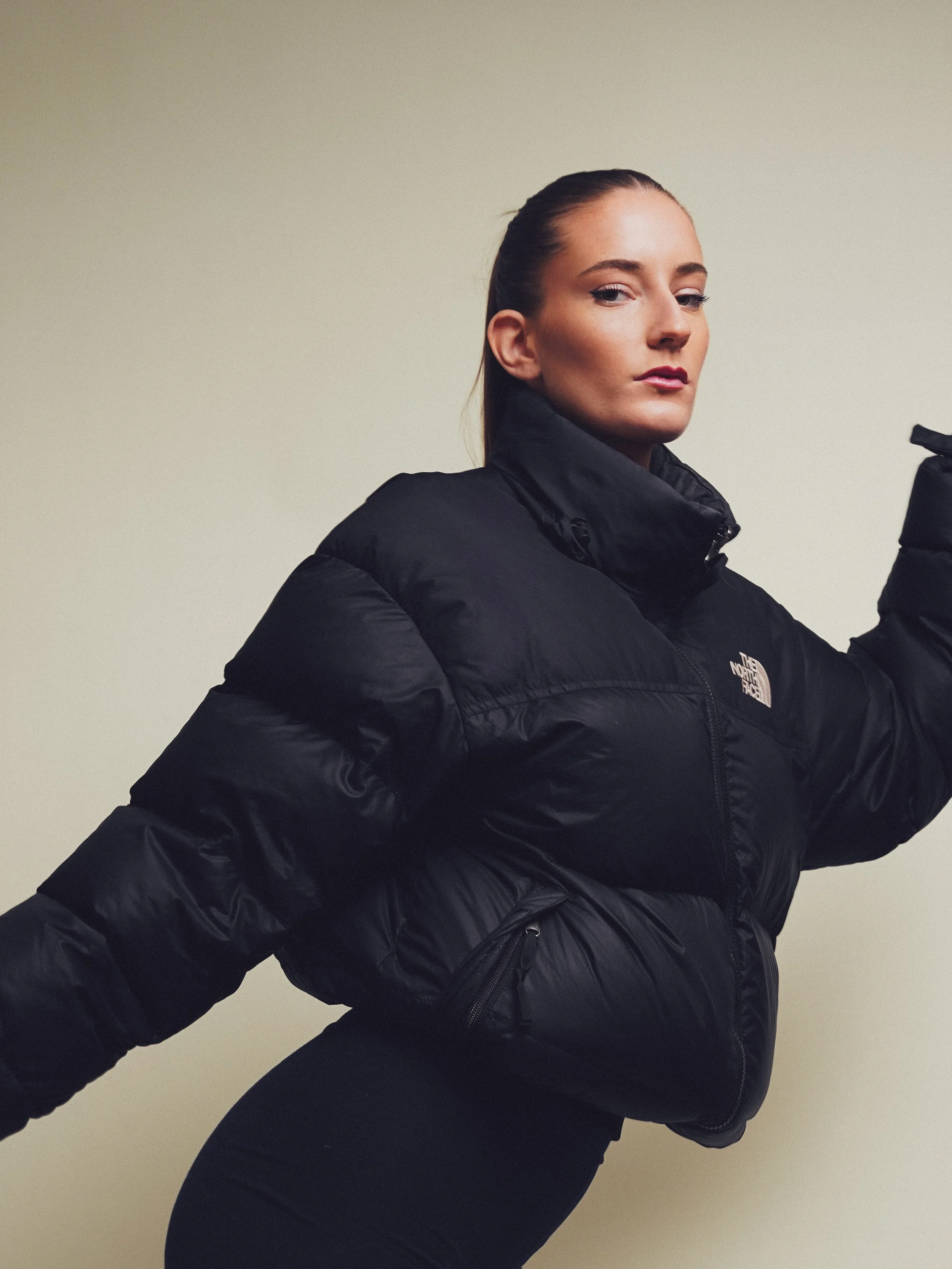 A woman in a black North Face puffy jacket with tied-back hair, standing sideways against a plain beige wall.