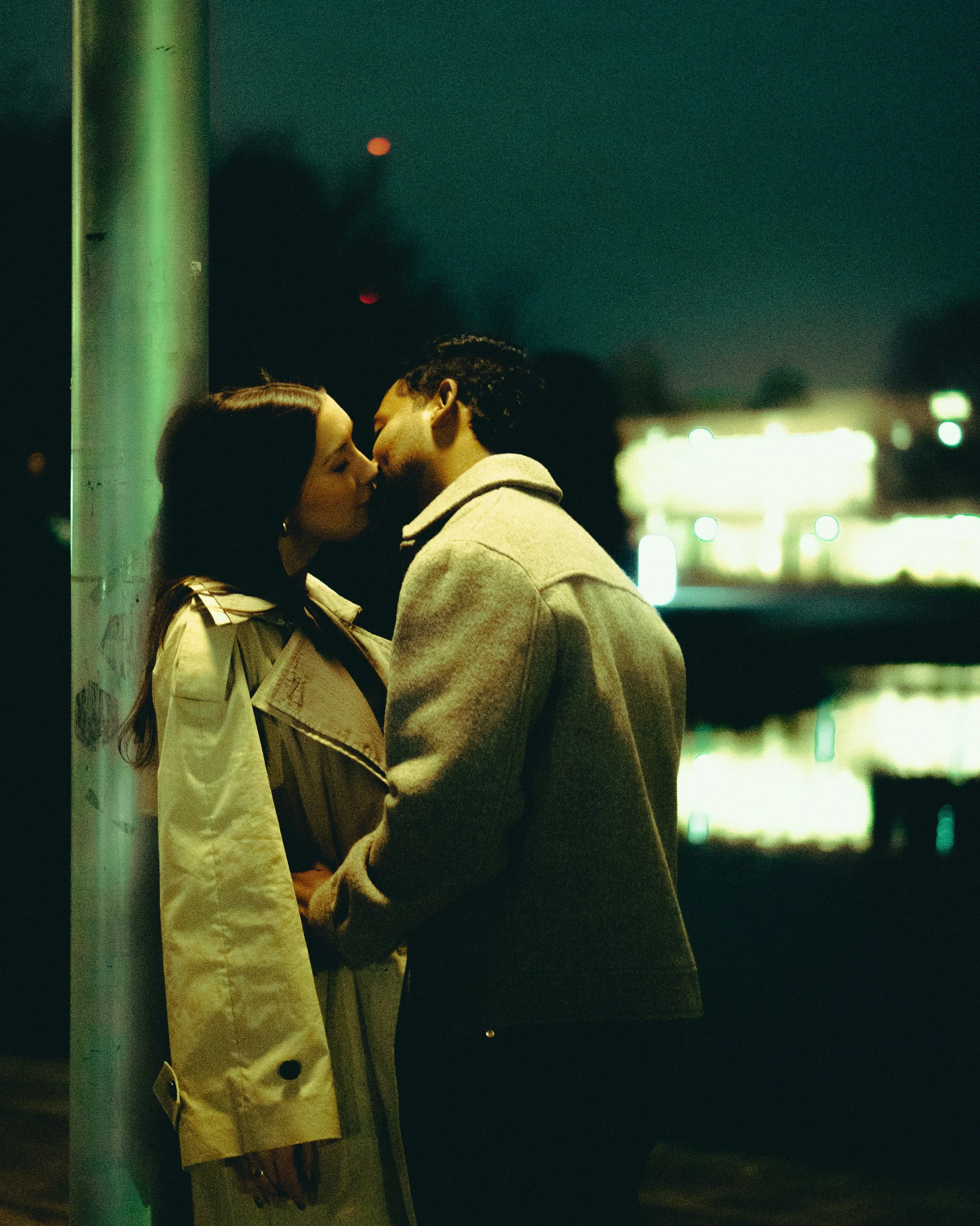 A couple sharing a kiss at night near a light pole, with blurred city lights in the background.
