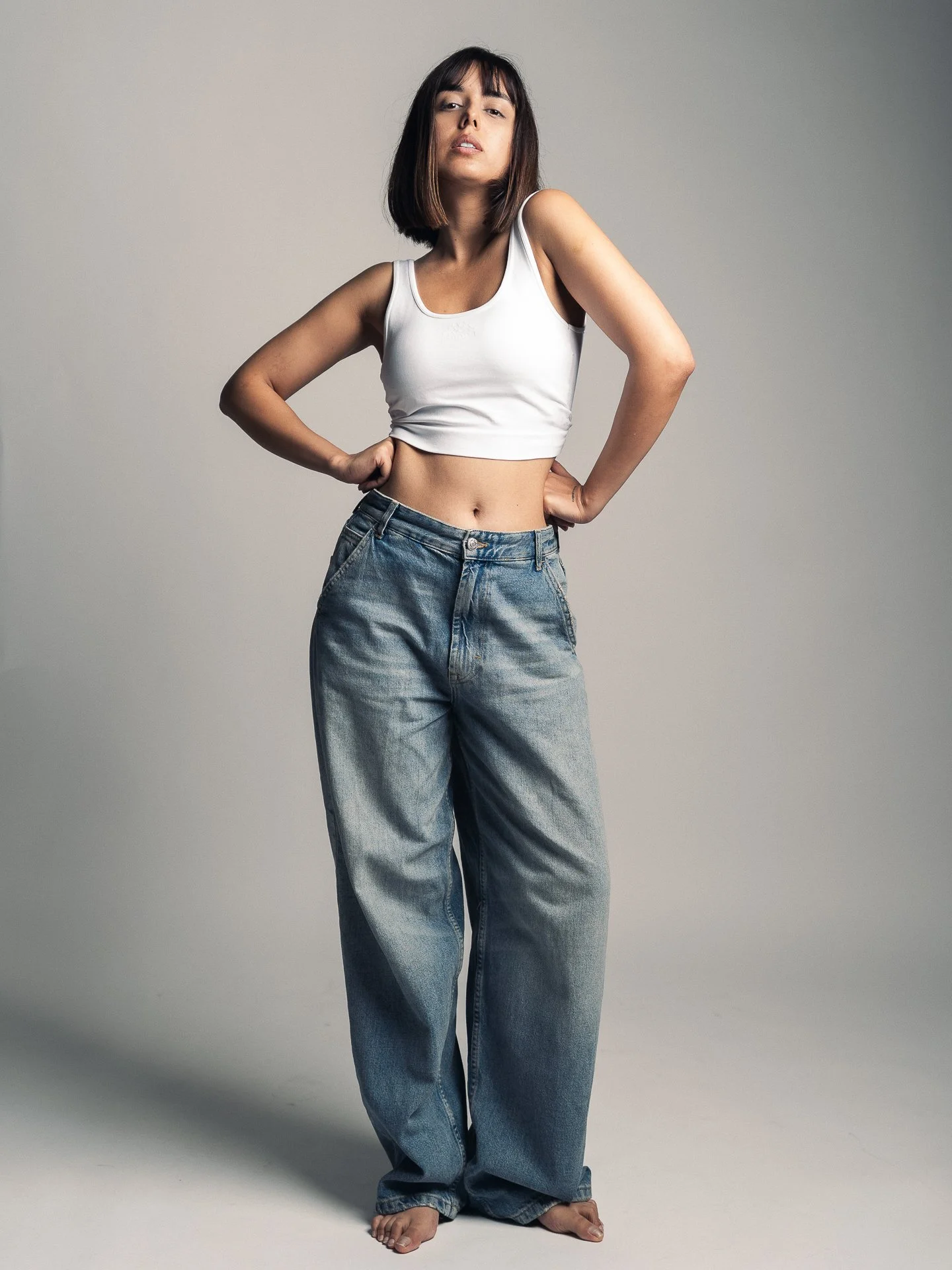 A woman with shoulder-length dark hair wearing a white crop top and baggy jeans, standing against a plain gray background.