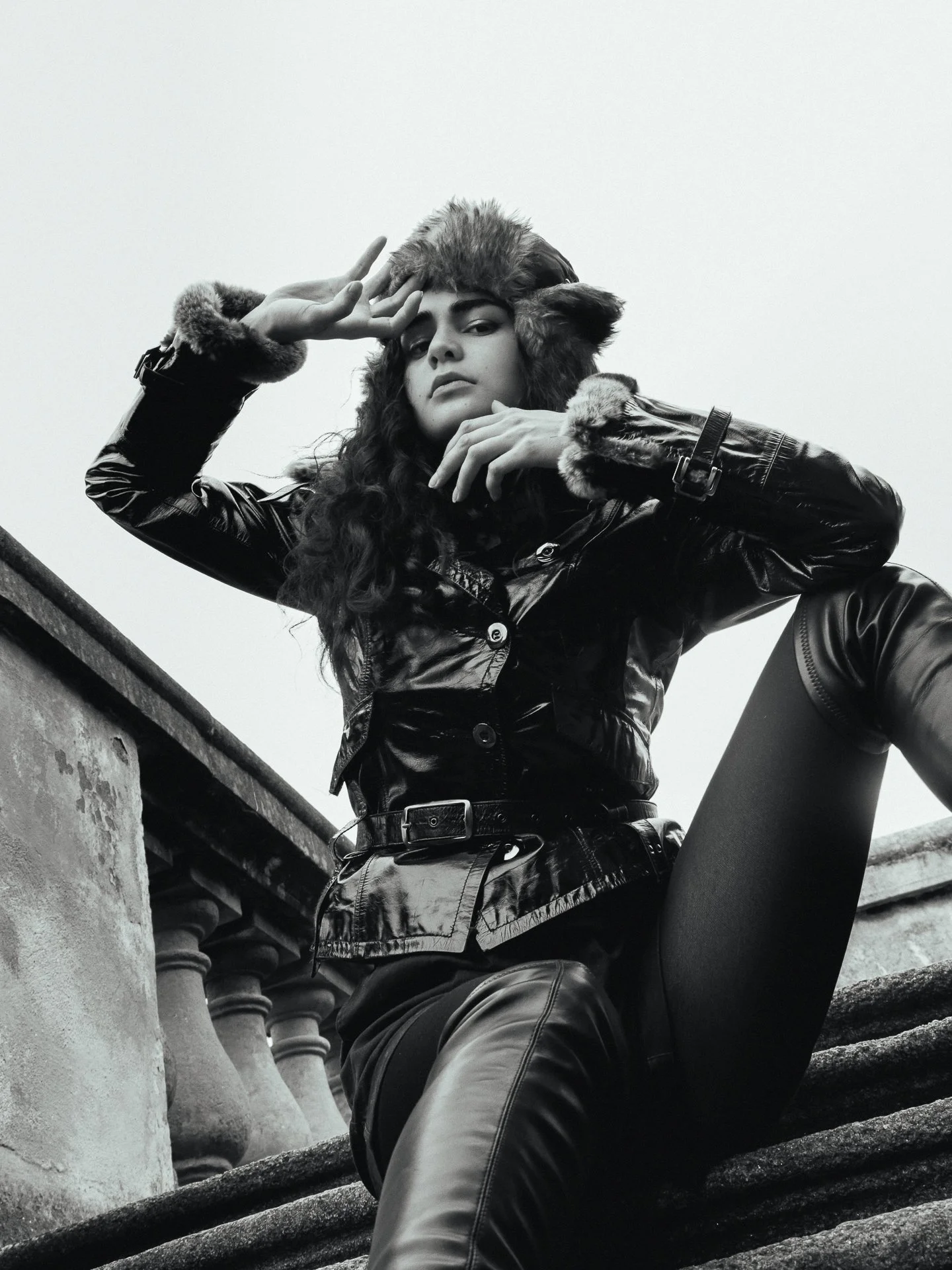 A woman wearing a fur-trimmed hat and a shiny leather jacket, sitting on stairs with her leg raised, looking down at the camera in a black-and-white photograph.