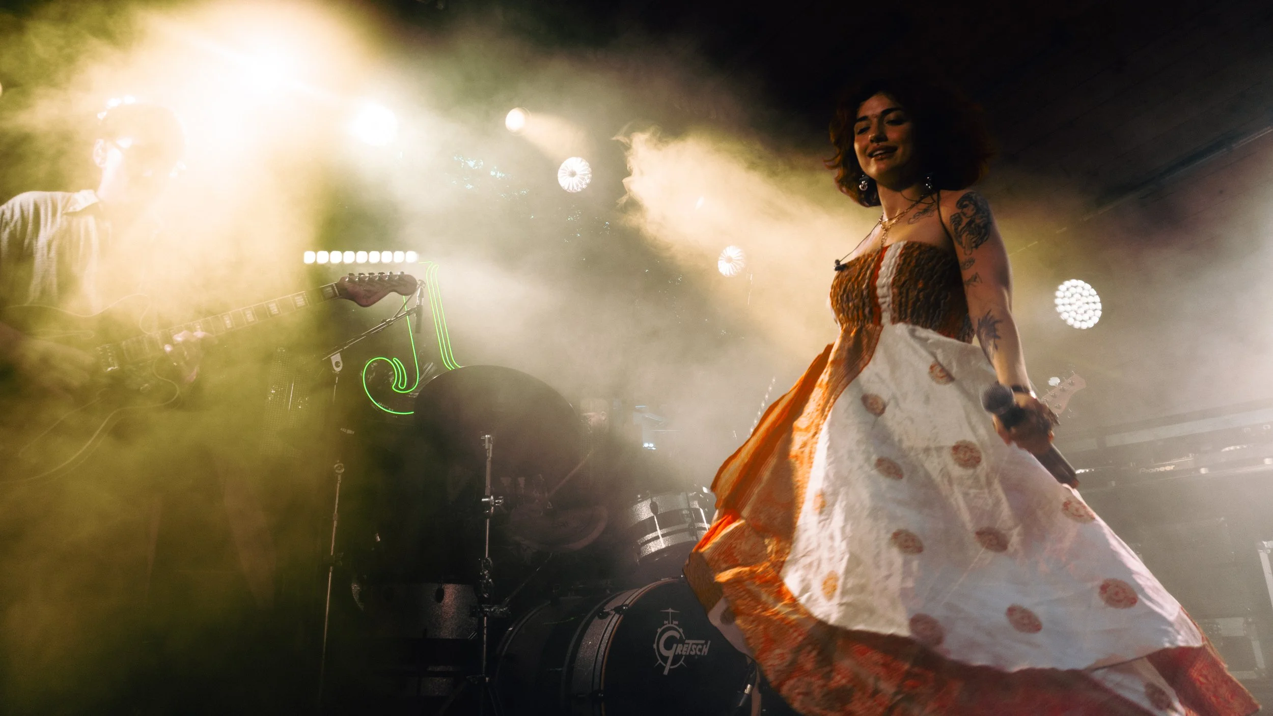A woman with curly hair, tattoos, and wearing a colorful dress performing on stage with a microphone, with a musician partially visible playing guitar behind her and stage lights creating a smoky, vibrant atmosphere.