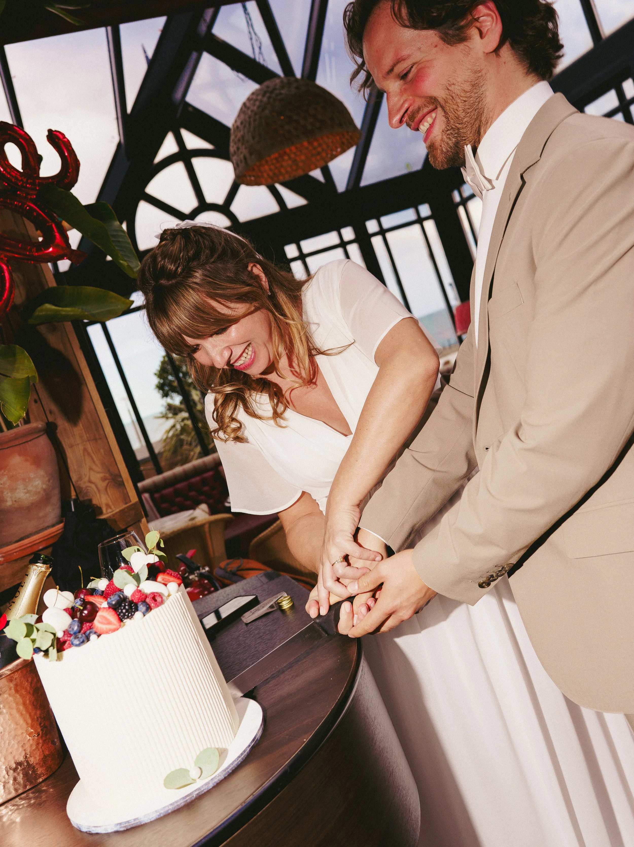A happily smiling woman and man in a beige suit are cutting a wedding cake together in a bright, modern room with large windows and decorative ceiling beams.