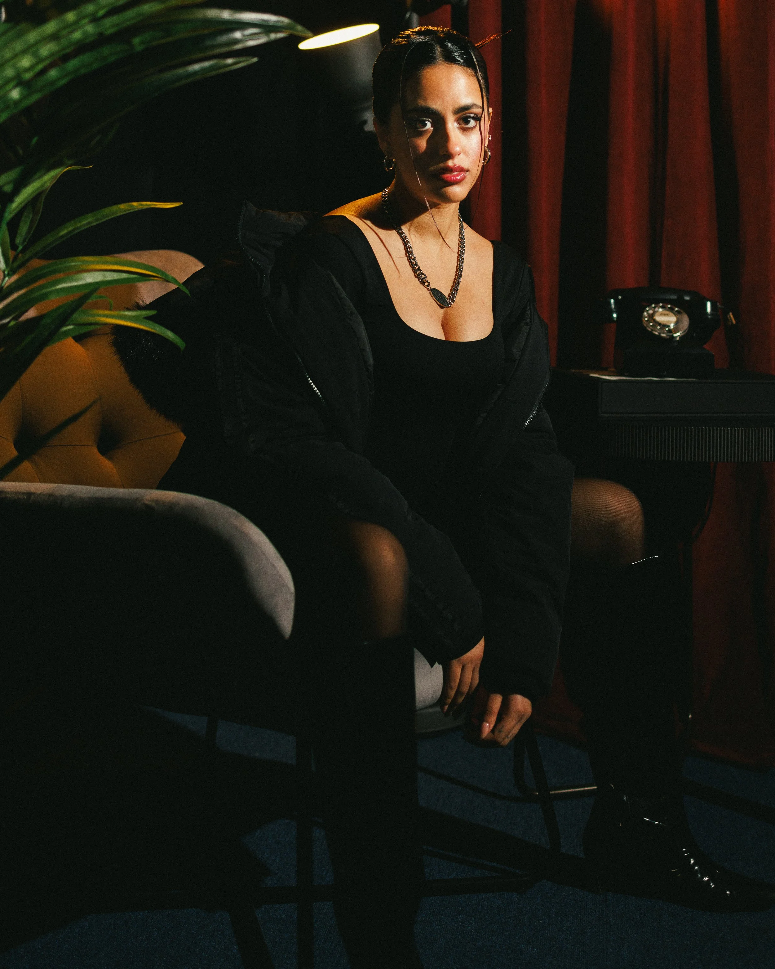 A woman sitting on a chair in a dimly lit room with plants, red curtains, and an old-fashioned rotary phone on a table, wearing a black dress and jewelry.