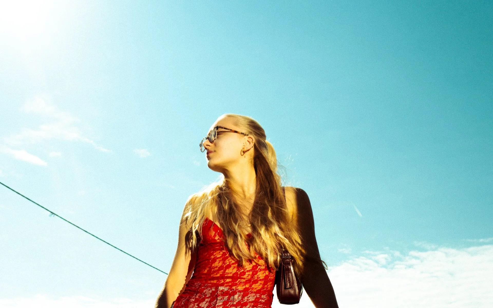A woman with long blonde hair wearing stylish sunglasses and a red lace dress, standing outdoors against a clear blue sky.