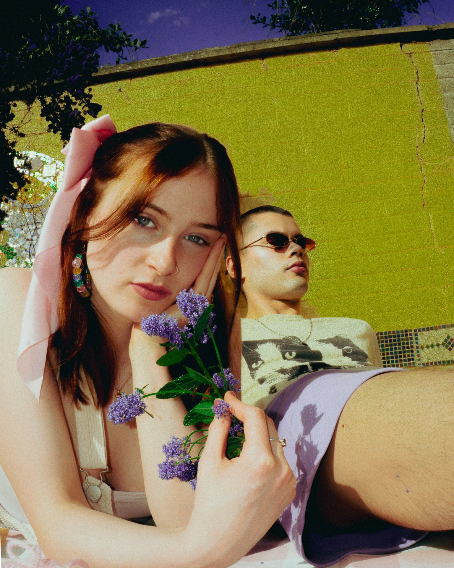 A young woman with red hair, wearing a white headband with a pink bow, and a young man with short hair and sunglasses, relaxing outdoors against a green wall with a purple sky in the background. The woman is holding purple flowers and resting her hea