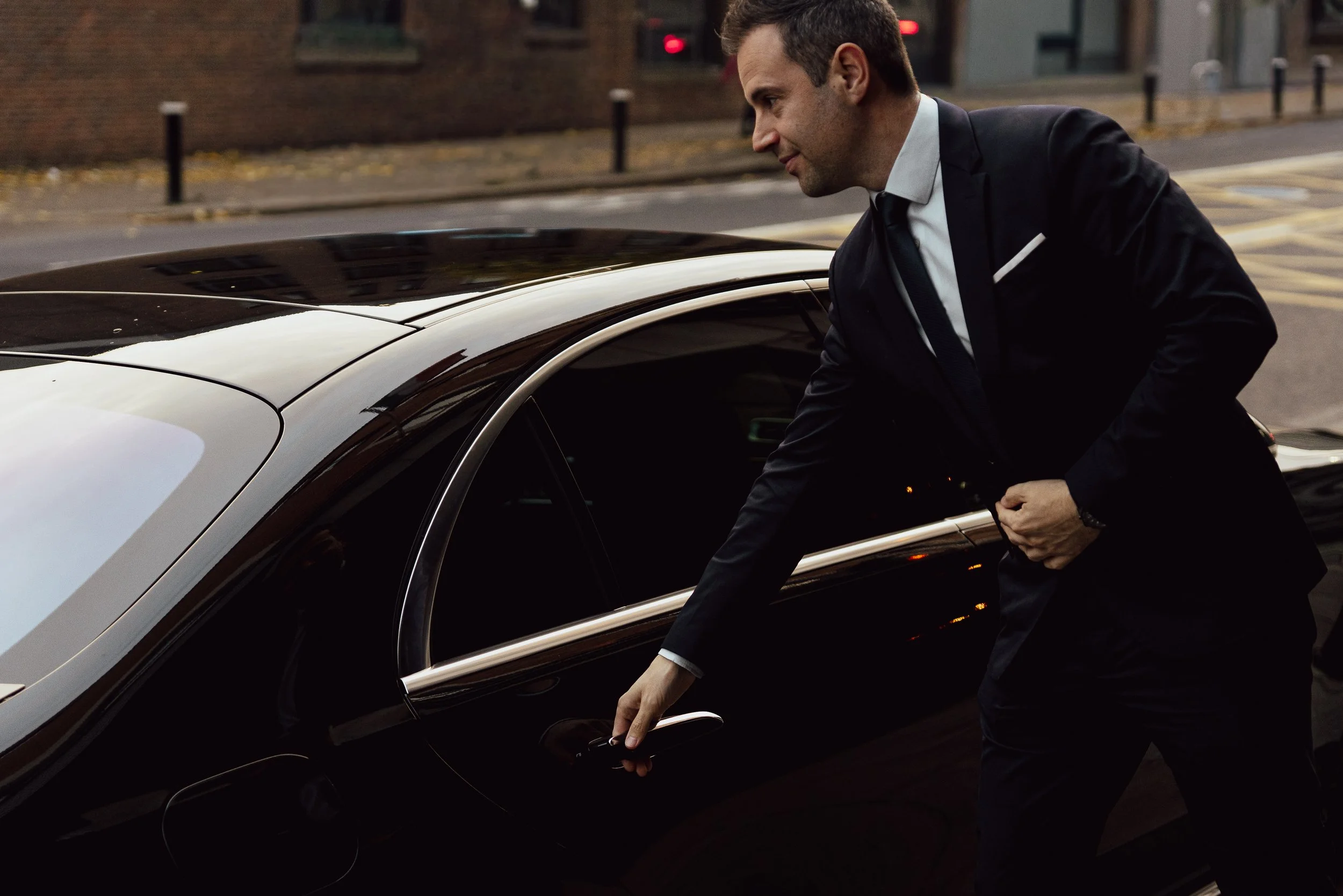 Man in a black suit opening the door of a black luxury car on a city street in the evening.