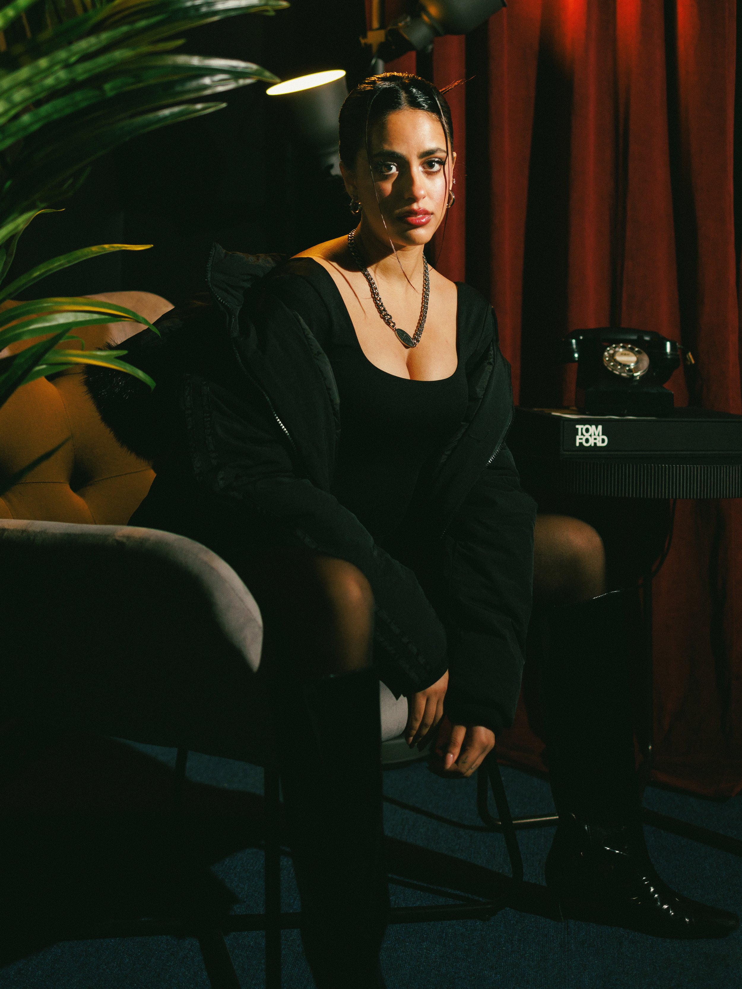A woman sitting on a chair in a dimly lit room with red curtains, wearing a black outfit and jewelry, with a vintage rotary phone on a table nearby.