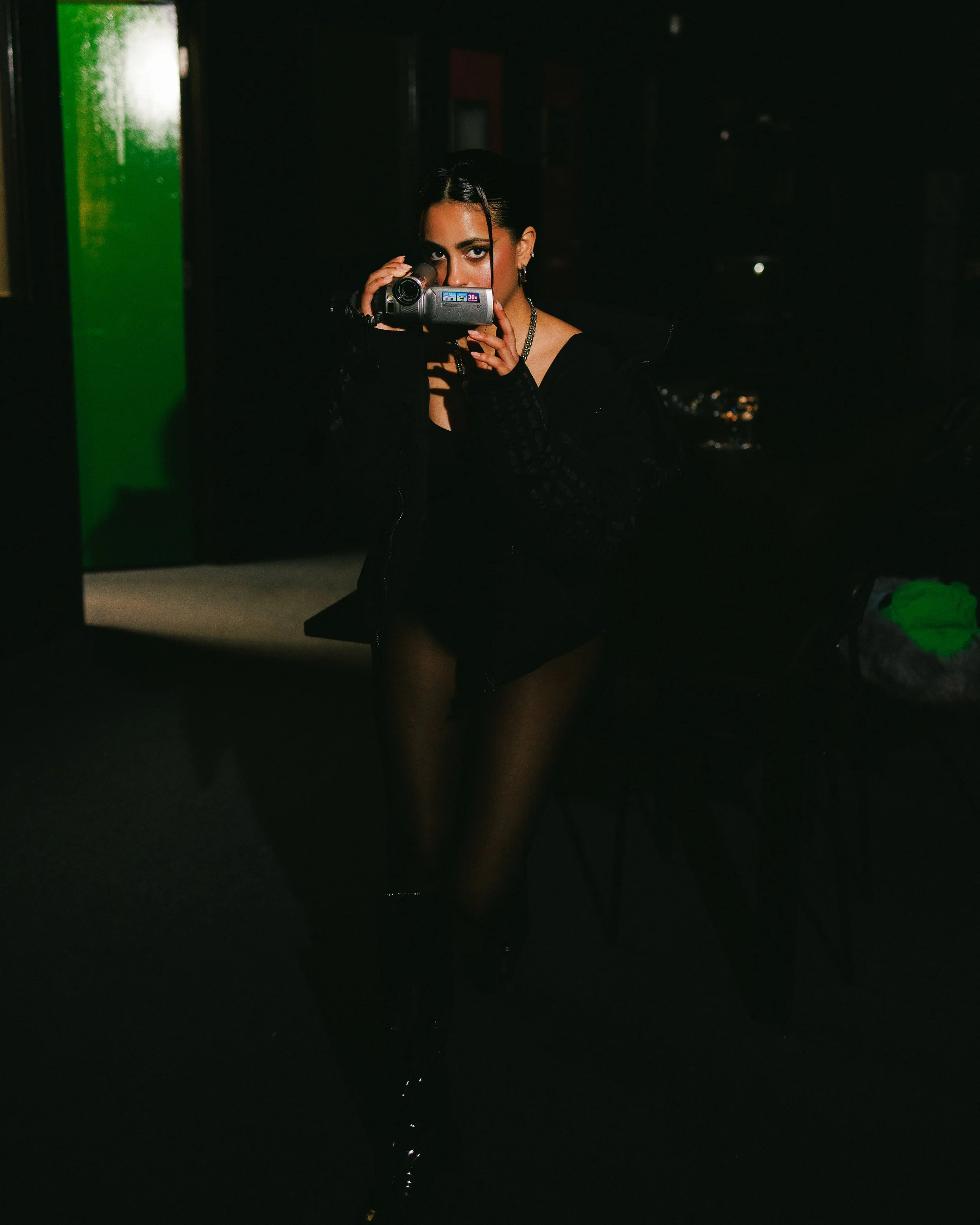A woman with dark hair taking a photo with a camcorder in a dimly lit indoor setting, wearing black clothing and jewelry.