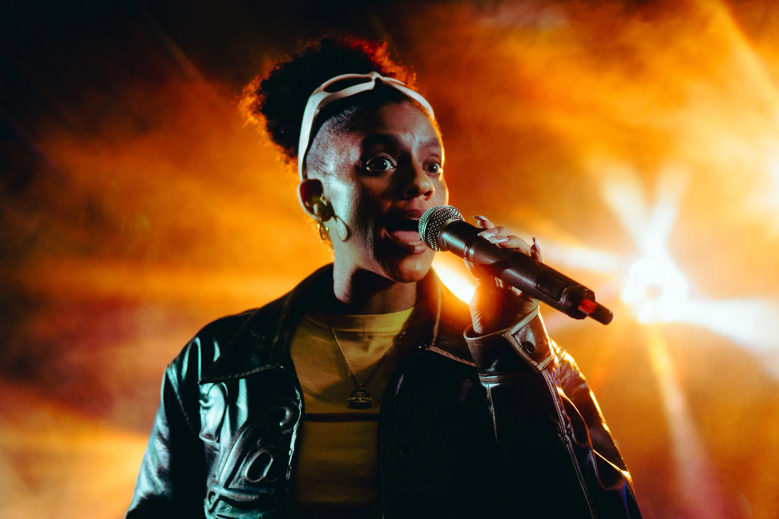 A woman singing into a microphone on stage with bright orange lighting in the background.