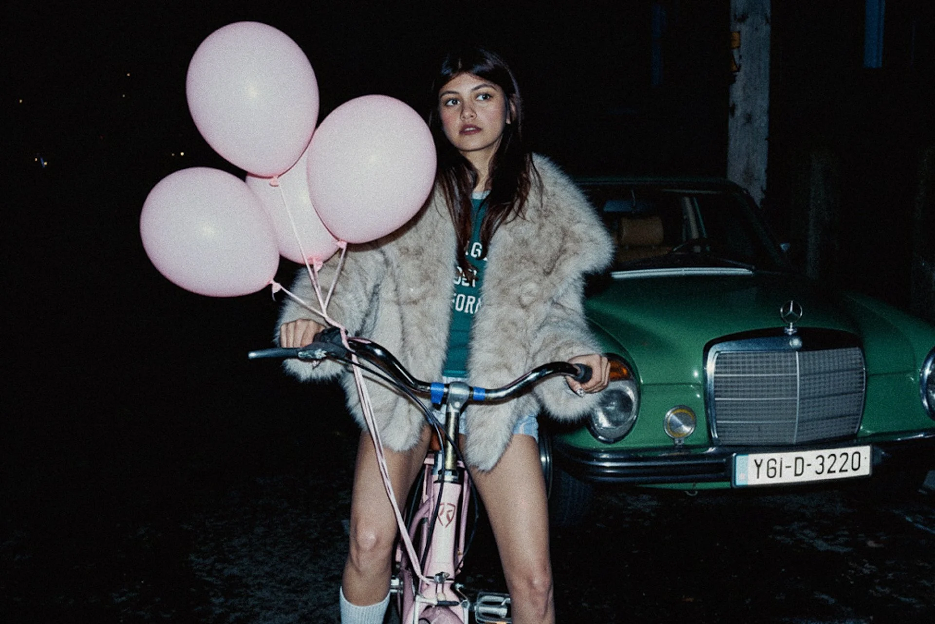 A young woman with dark hair and fair skin standing at night, holding pink balloons and riding a pink bicycle, wearing a fur coat and shorts, with a dark vintage car in the background.