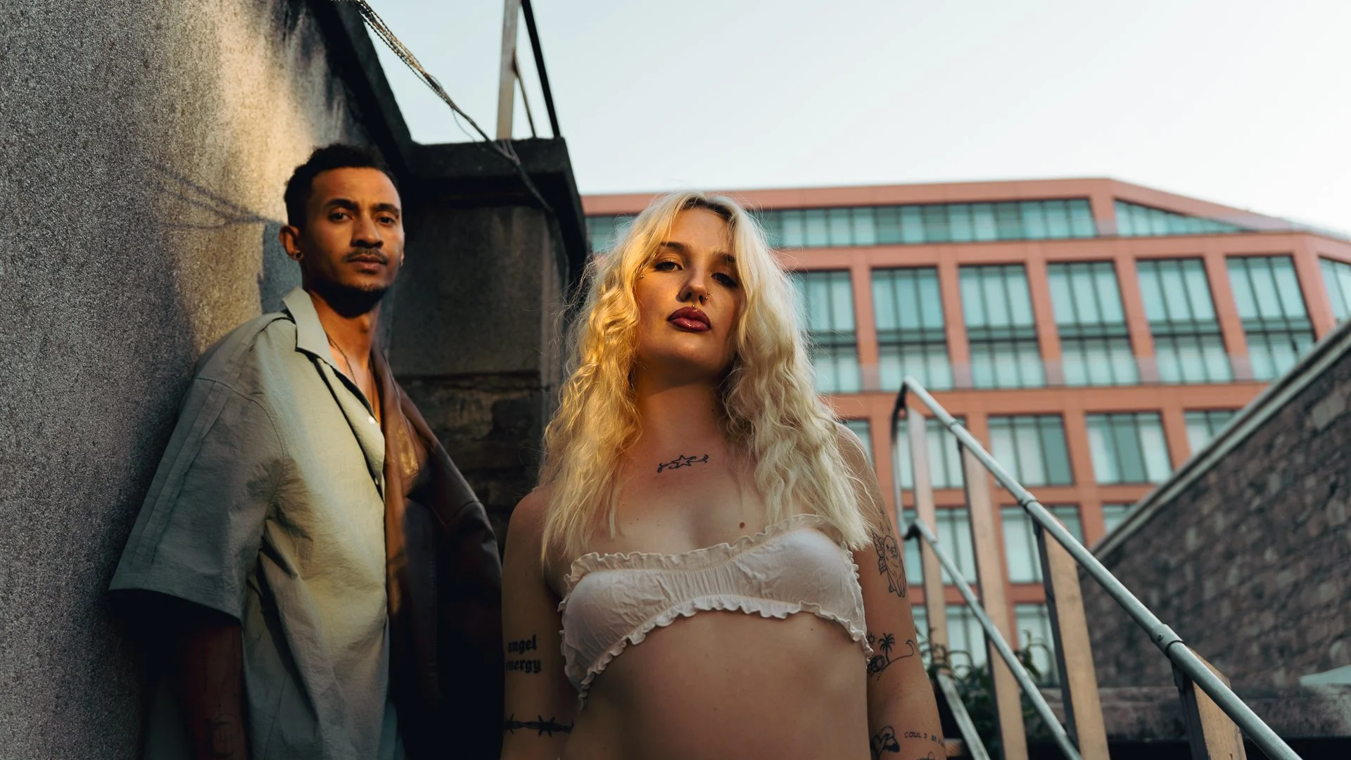 Two young adults, a man and a woman, standing on outdoor stairs near an urban building, with the woman in the foreground and the man in the background, both looking serious.