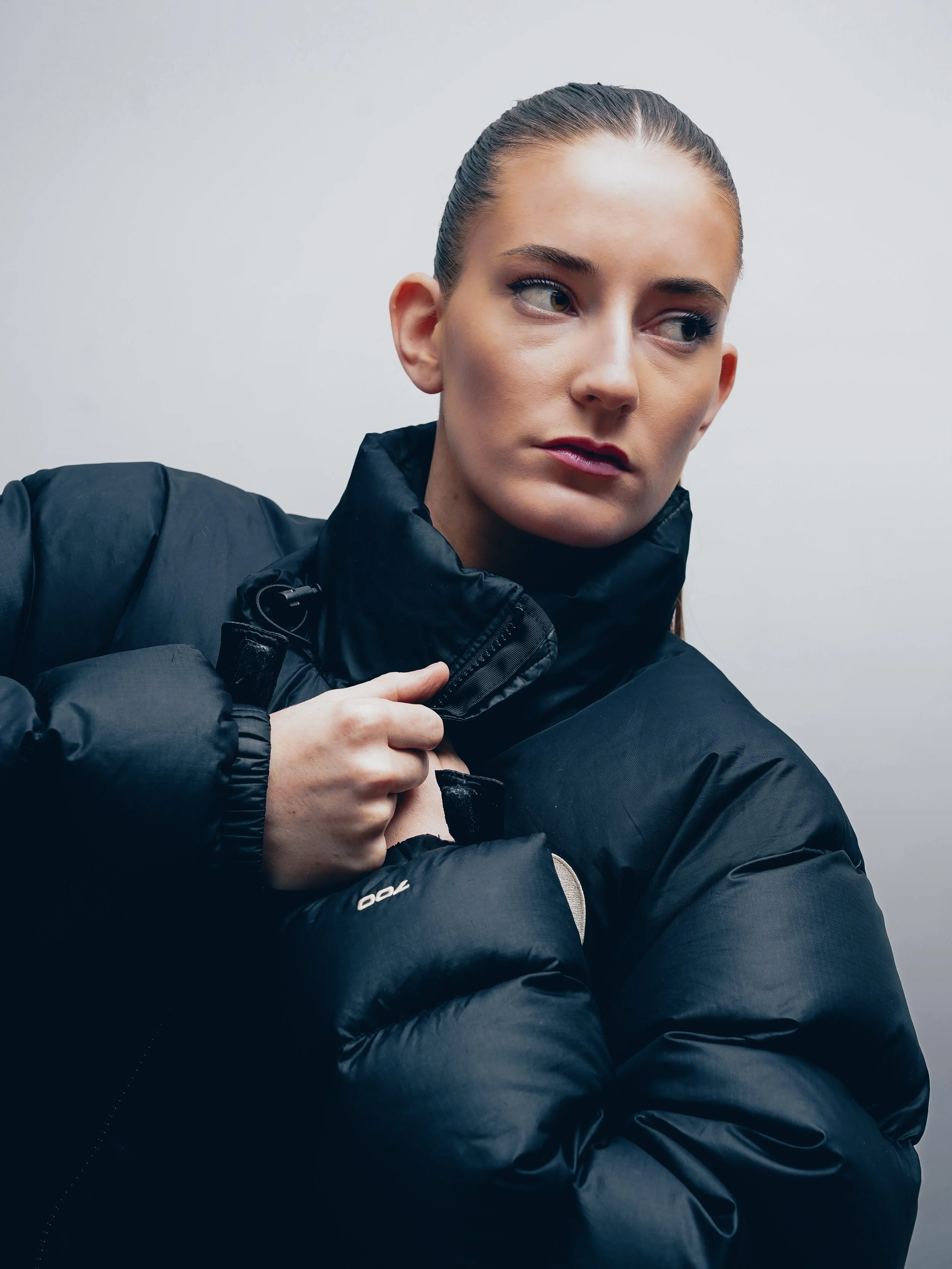 A woman with fair skin and brown hair pulled back, wearing a black puffer jacket, looking to her right with a serious expression.