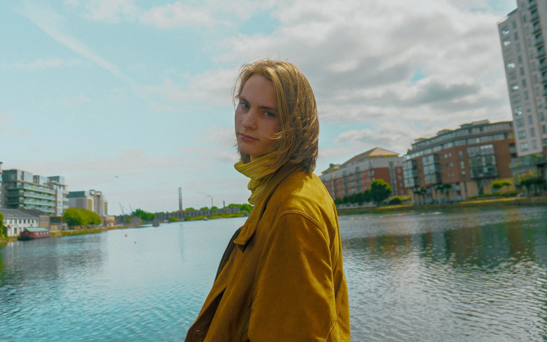 A young woman with shoulder-length hair and a yellow jacket stands by a river in an urban area with modern buildings, under a partly cloudy sky.