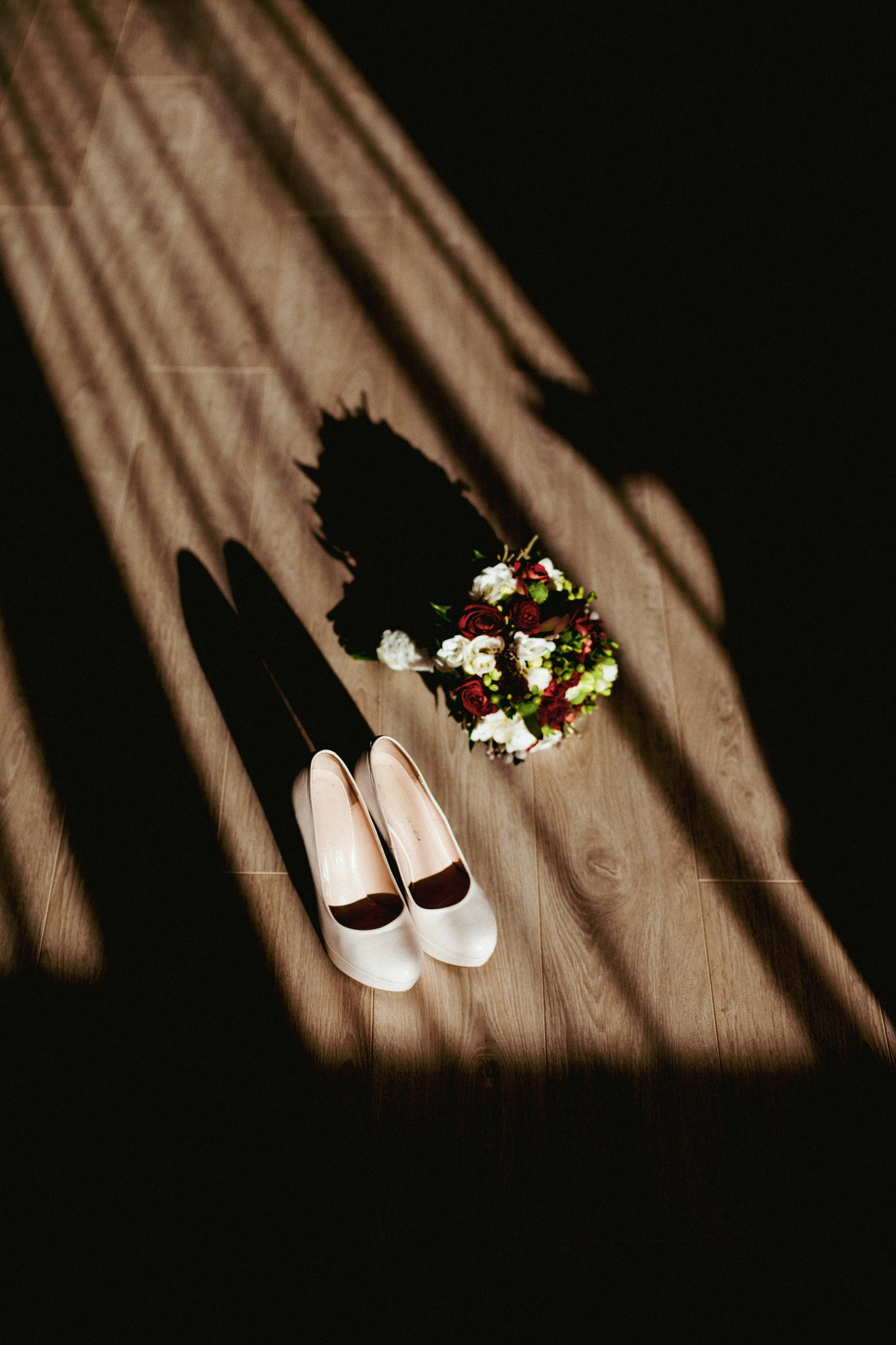 A pair of white high heels and a bouquet of flowers cast a shadow on a wooden floor, seen through a shadow of a chair.