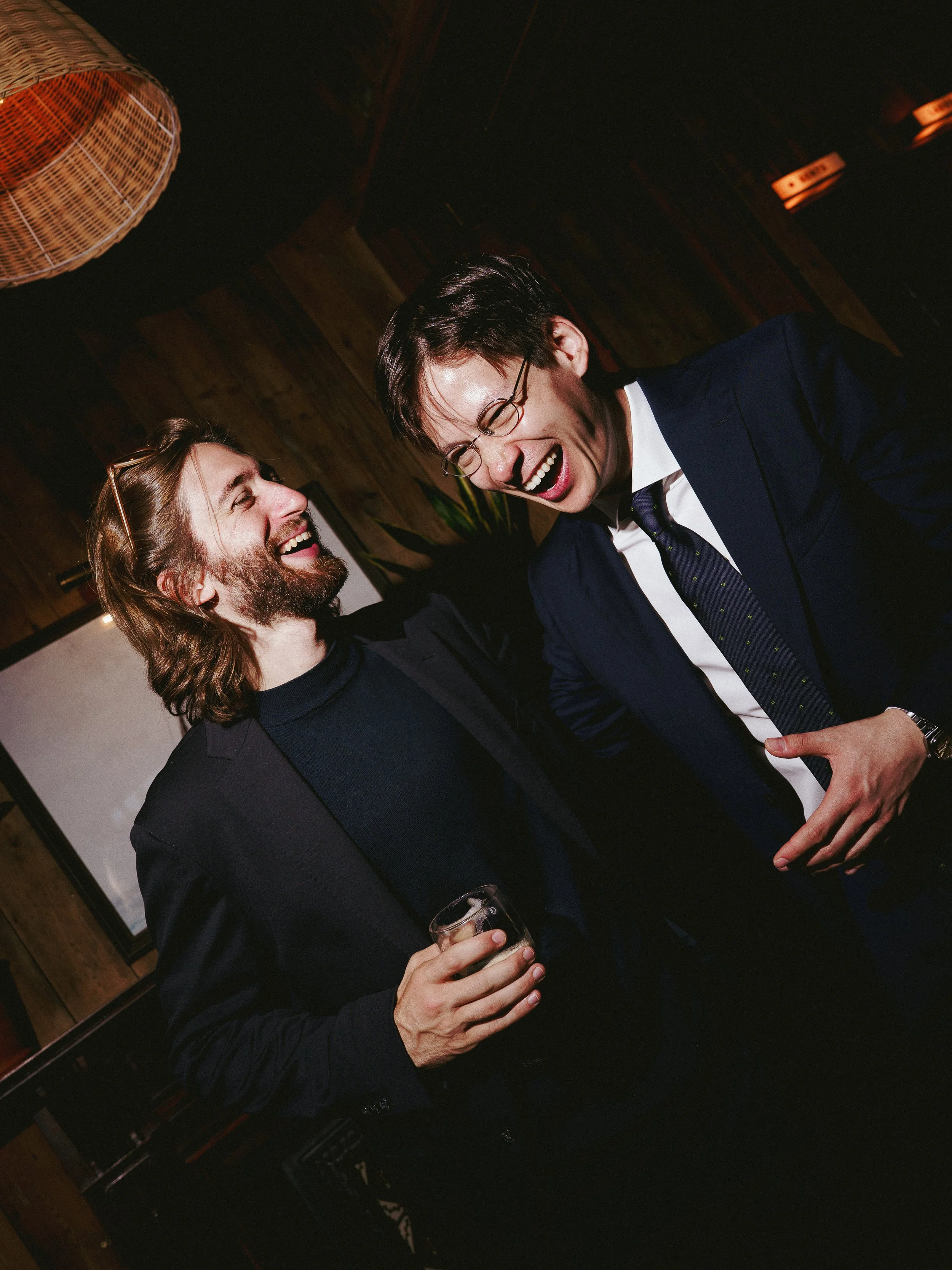 Two men are laughing and talking together at a social event. One man is holding a drink, and both appear to be having a good time.