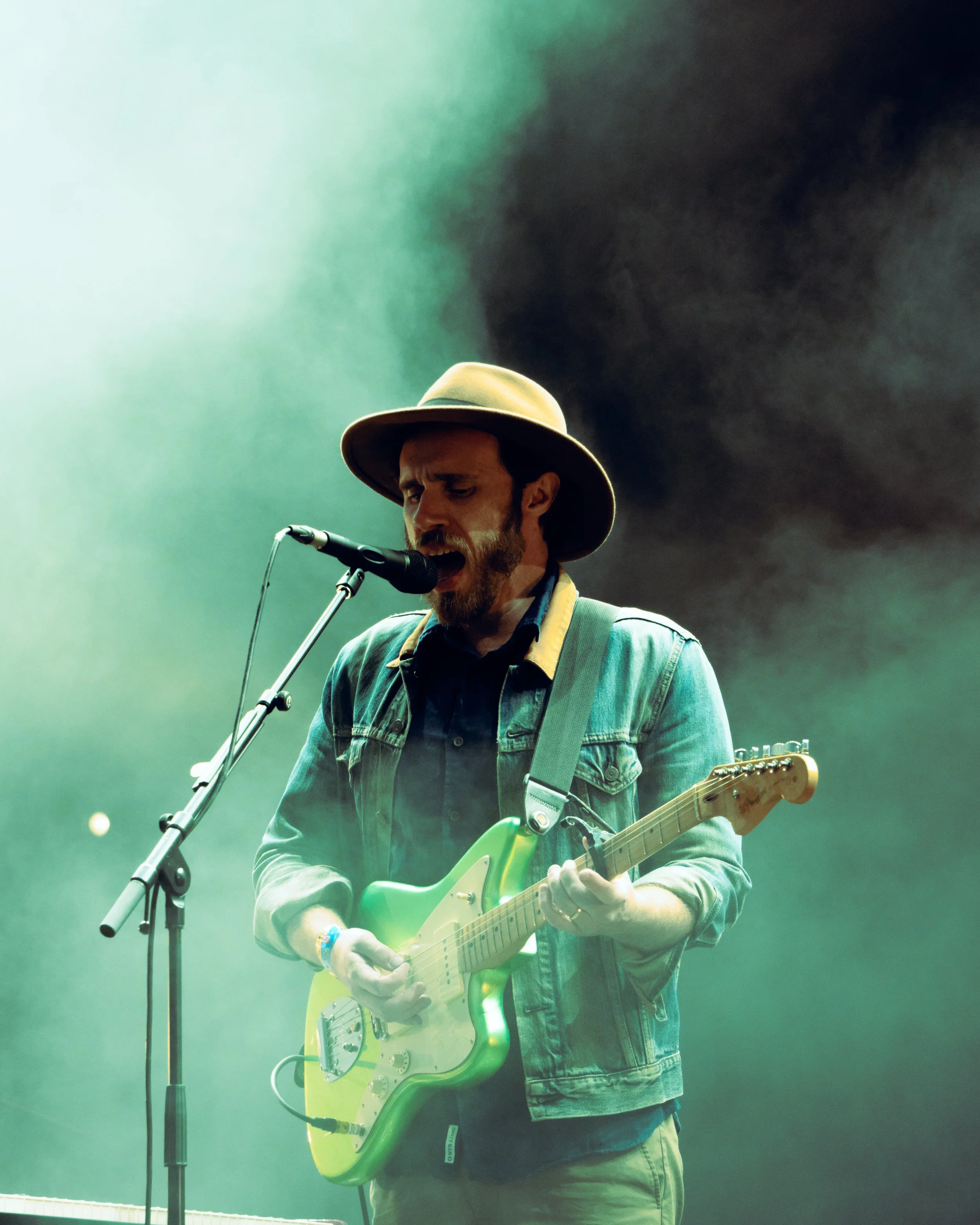A man singing and playing an electric guitar on stage, wearing a hat and denim jacket, surrounded by green stage lighting and fog.