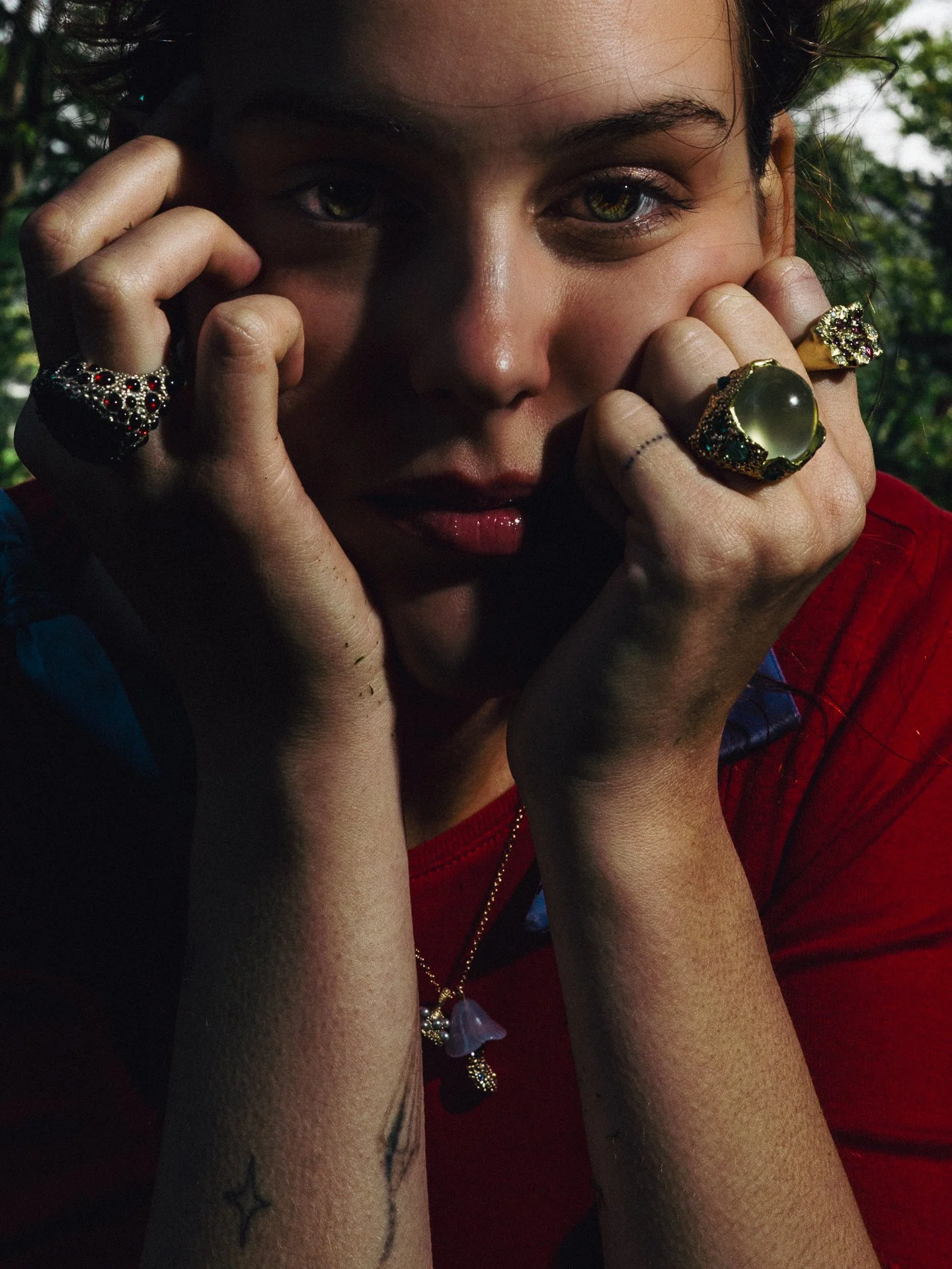 Close-up of a woman with short dark hair, wearing multiple rings and a necklace with a purple heart-shaped pendant. She holds her face with her hands, her fingers near her eyes. She is wearing a red top and has a small tattoo of stars on her forearm. The background is outdoors with green foliage.