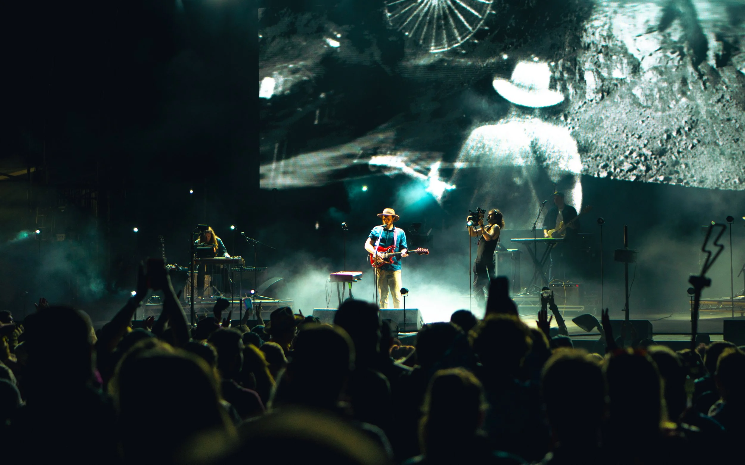 Live music concert with a band playing on stage, a large crowd in front, a performer with a guitar wearing a hat, and a black-and-white projected image of a person in the background.