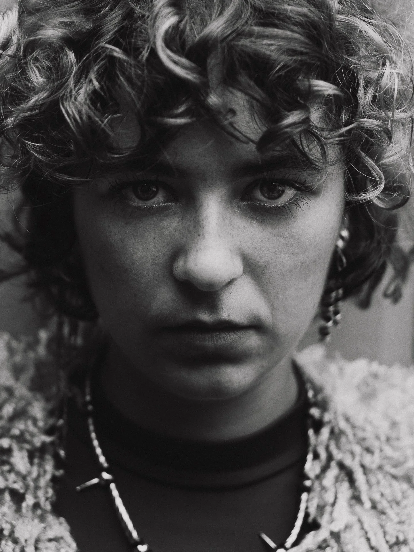 Close-up black and white portrait of a woman with curly hair, freckles, and intense expression, wearing a necklace and earrings.