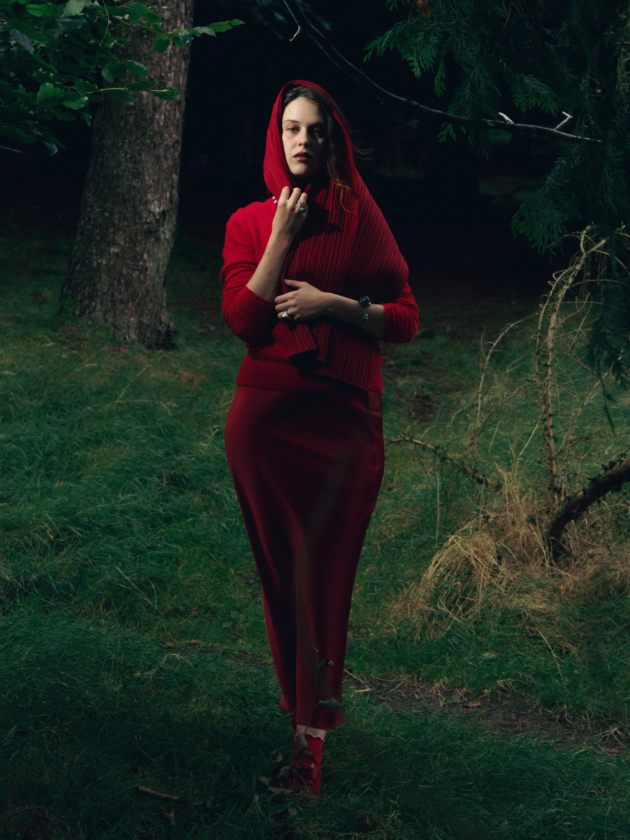 A woman in a red hoodie and matching long skirt stands in a nighttime forest, surrounded by trees and grass, with a serious expression.