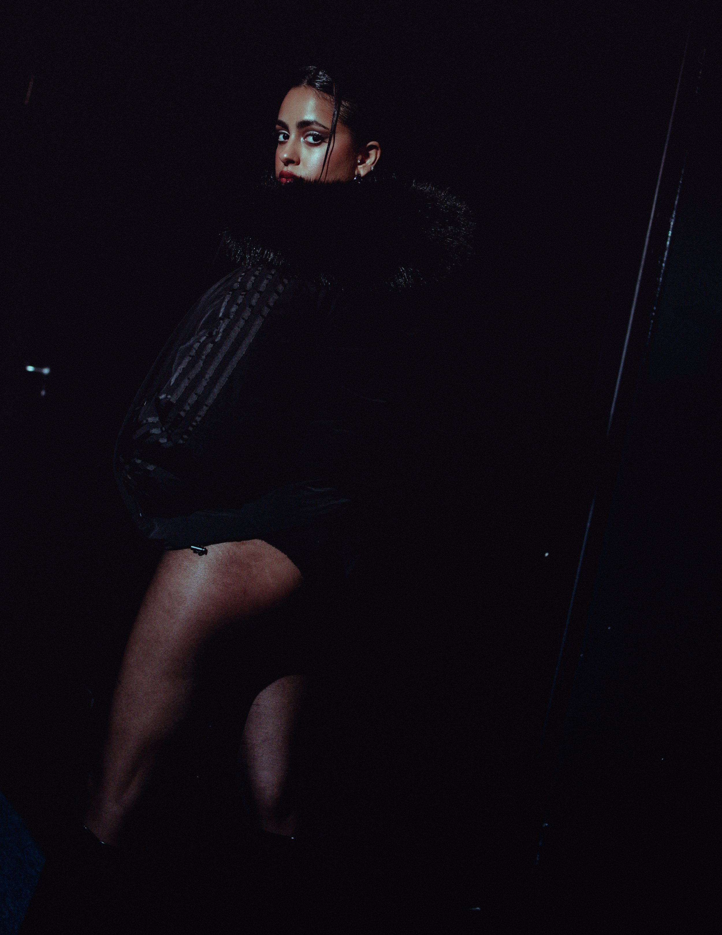 A woman in a black outfit with a fur collar, standing in a dark setting with minimal lighting, looking over her shoulder.
