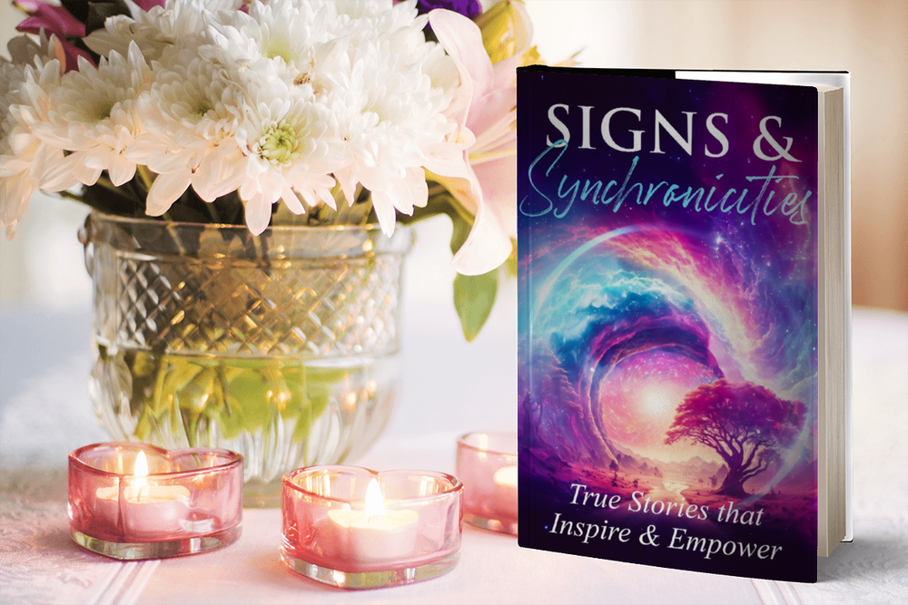 A book titled "Signs & Synchroncities" with a colorful cosmic and nature scene on the cover, placed next to a bouquet of white and pink flowers in a patterned glass vase and surrounded by small pink candles on a table.