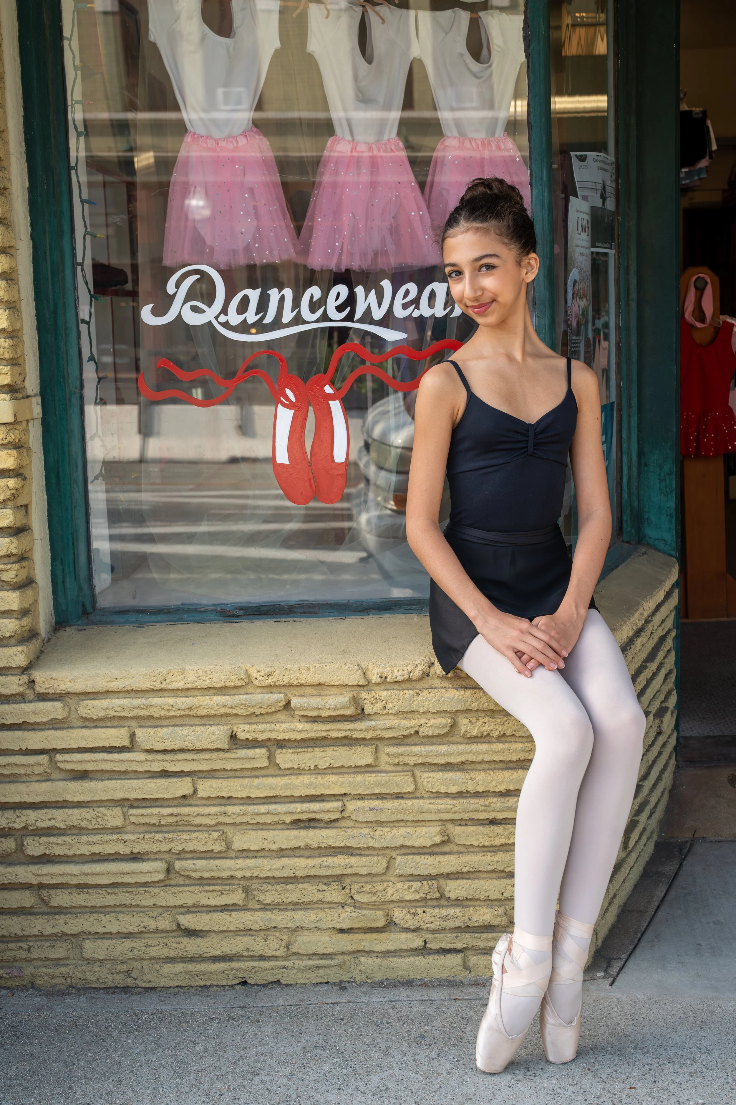 Young woman in ballet attire, sitting outside a dancewear shop with pink tutus and ballet slippers in the window, shop sign reading 'Dancewear' with a ballet slipper illustration.