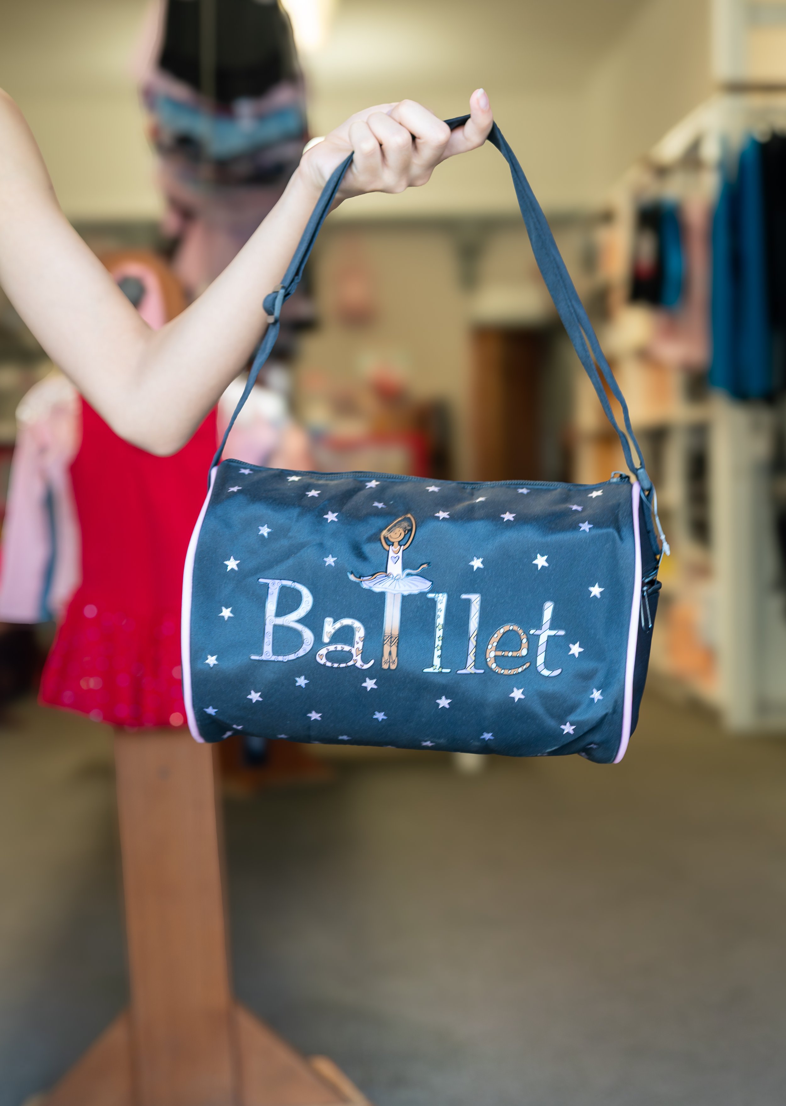 A child's navy blue bag with a ballerina graphic and the word 'Ballet' on it, being held by a hand inside a store.