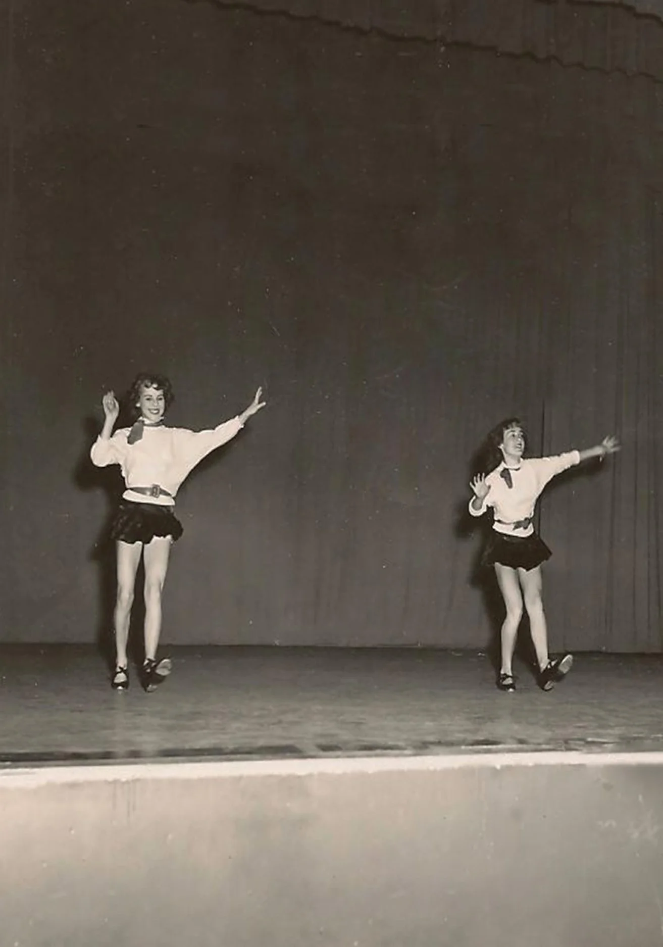 Black and white vintage photograph of two young girls performing a tap dance routine on a theater stage with curtain backdrop