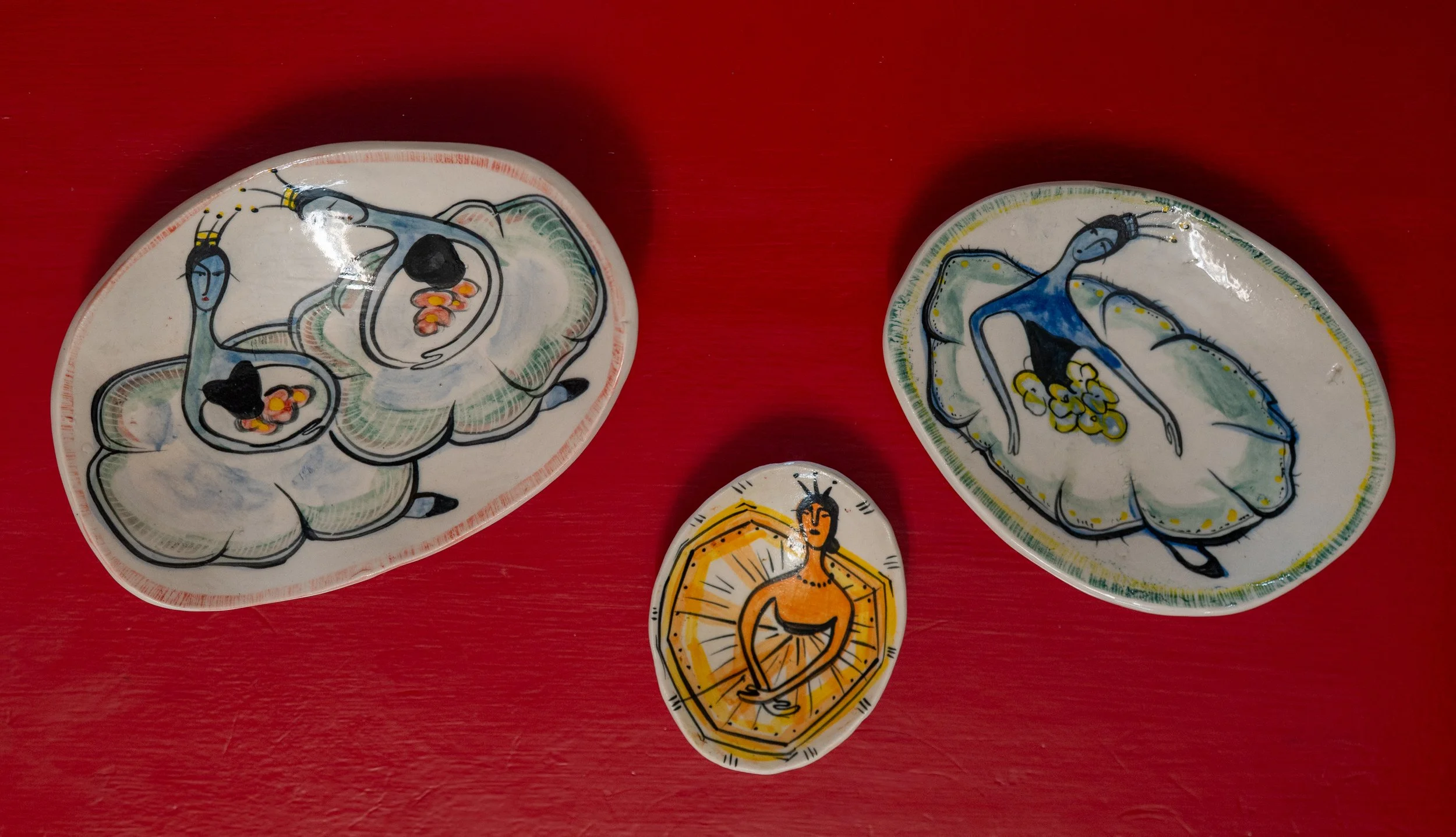 Decorative ceramic plates with abstract artistic illustrations on a red surface. Two large plates depict abstract figures, one in blue and one in gray, with floral elements. A smaller plate features a figure with a raised arm, surrounded by a radiati