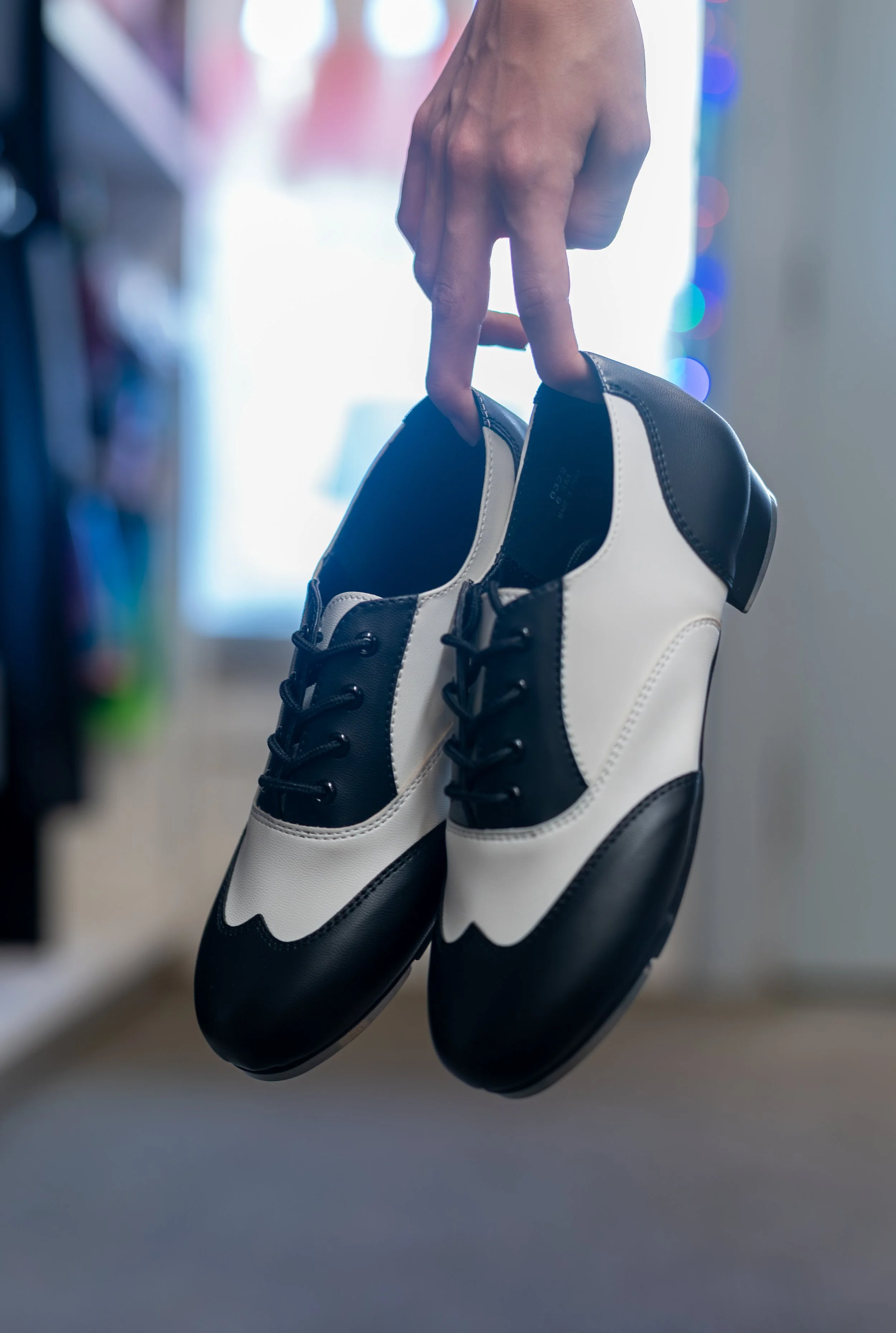 Person holding a pair of black and white tap shoes in a room with blurred background.