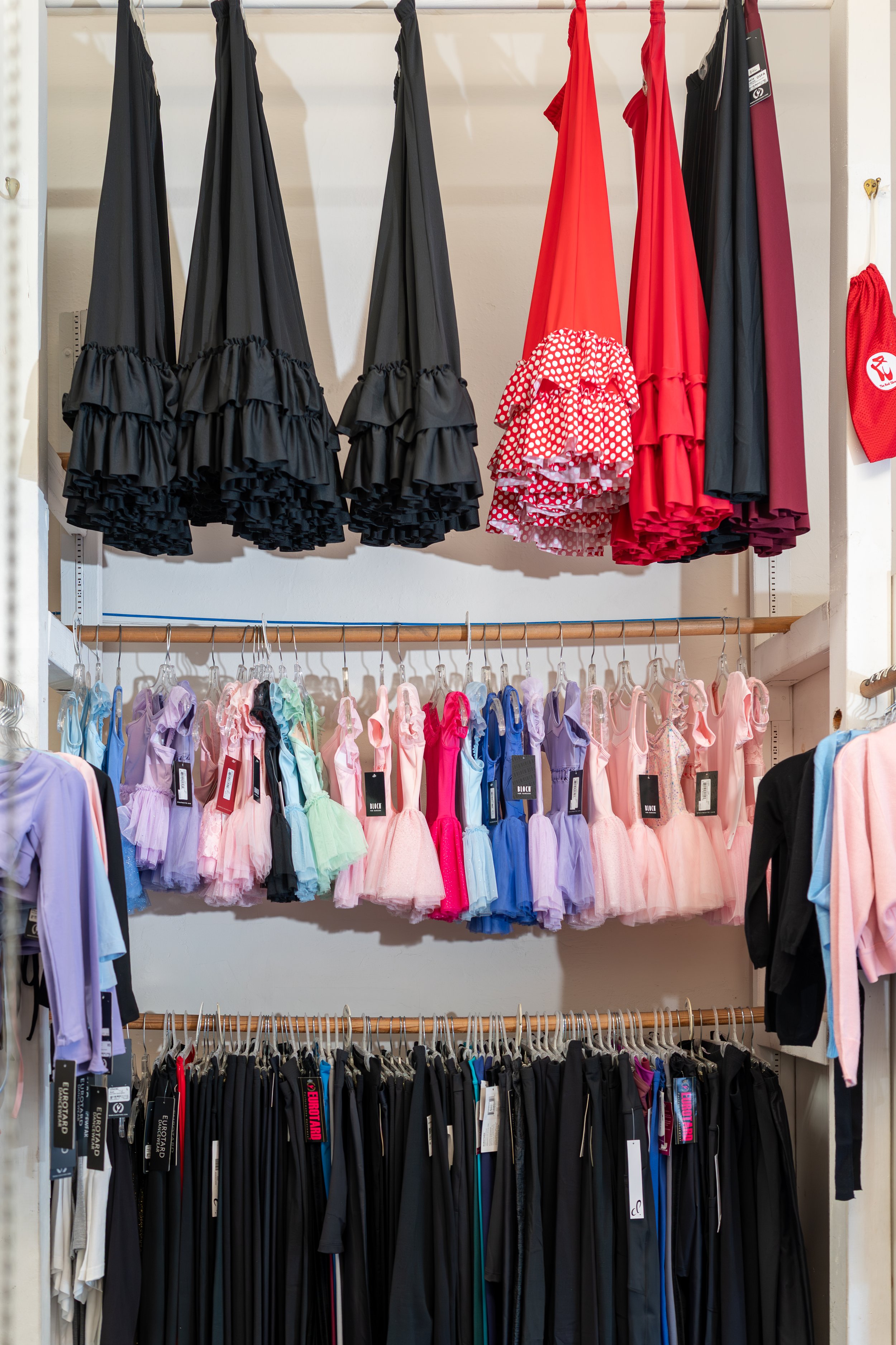 Clothing store display with black ruffled umbrellas hanging at the top, colorful children's dresses with ruffles and tags on the middle rack, and black and multicolored pants on racks below.