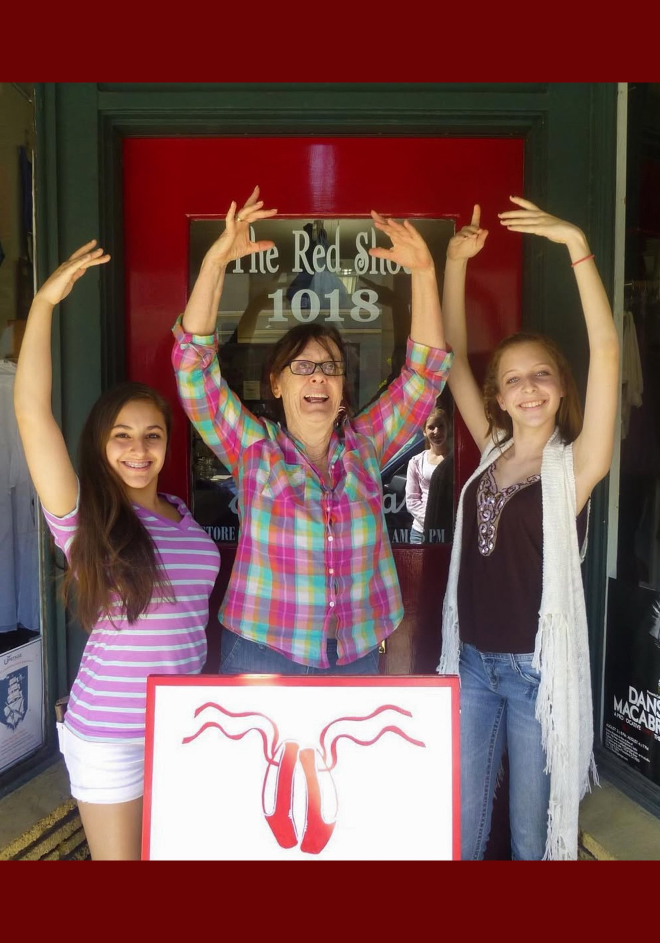 Three women standing outside a store, smiling with their arms raised above their heads. The woman in the middle is wearing a colorful plaid shirt and glasses, while the women on either side are in casual outfits. There is a sign in front of them with