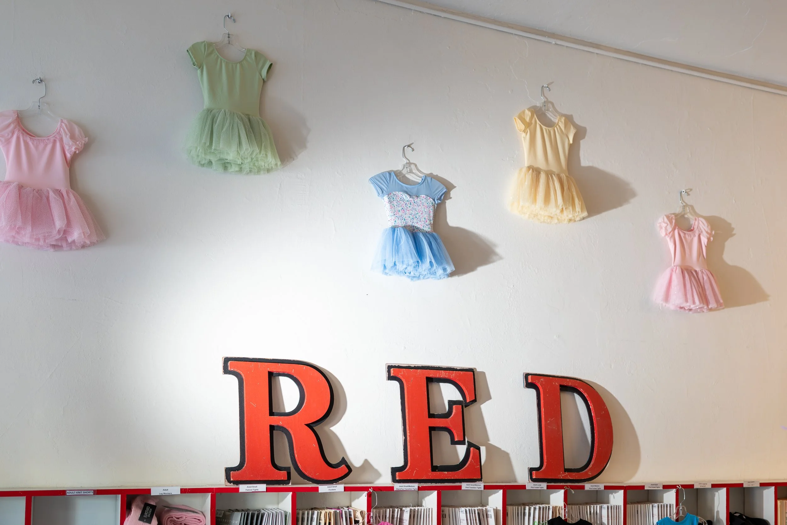 Colorful children's dresses hanging on a white wall above red shelving in a fabric store.