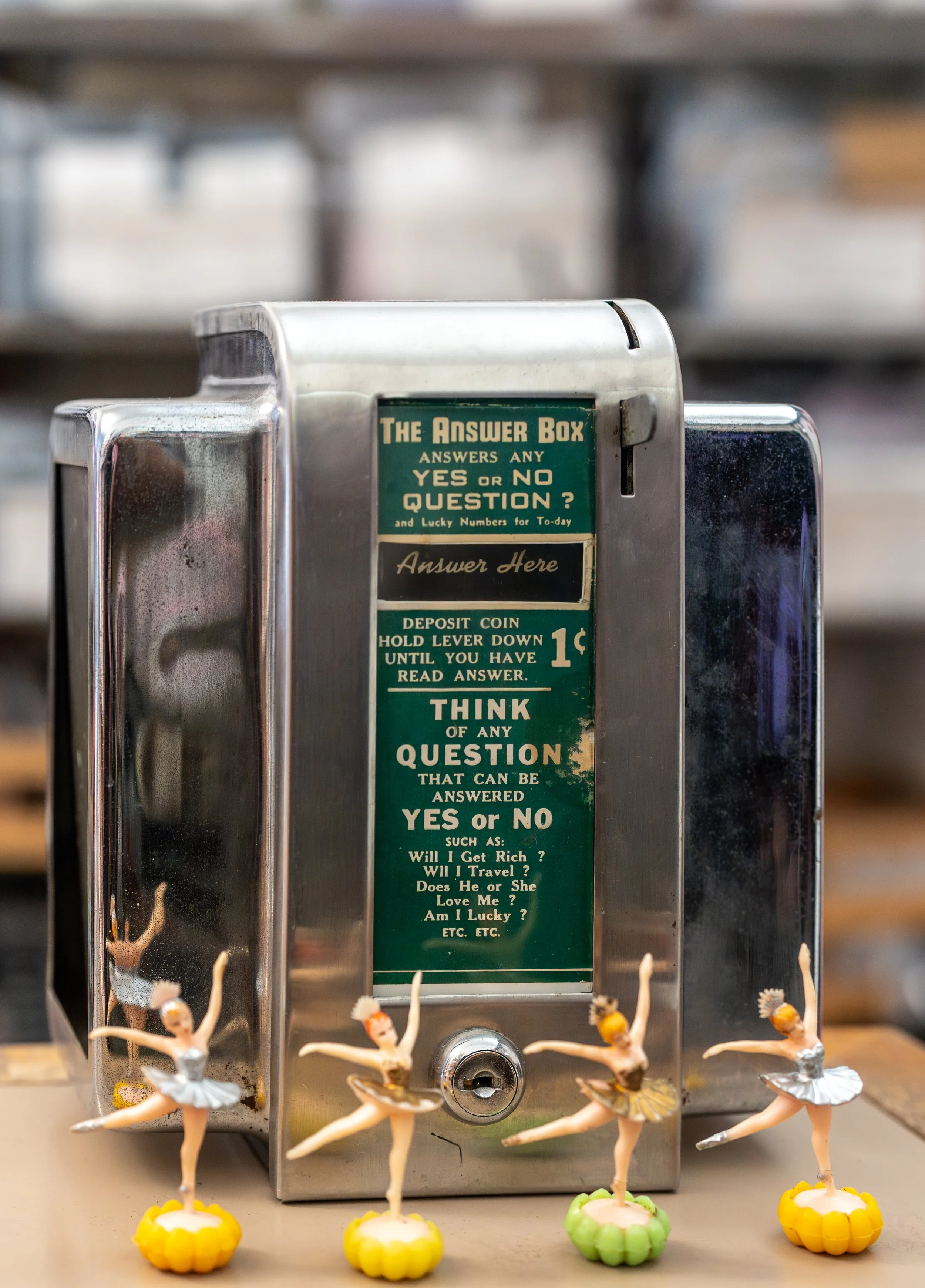 A vintage metal answer box with green and black sign instructions, decorated with small ballerina figurines arranged in a row in front of it.
