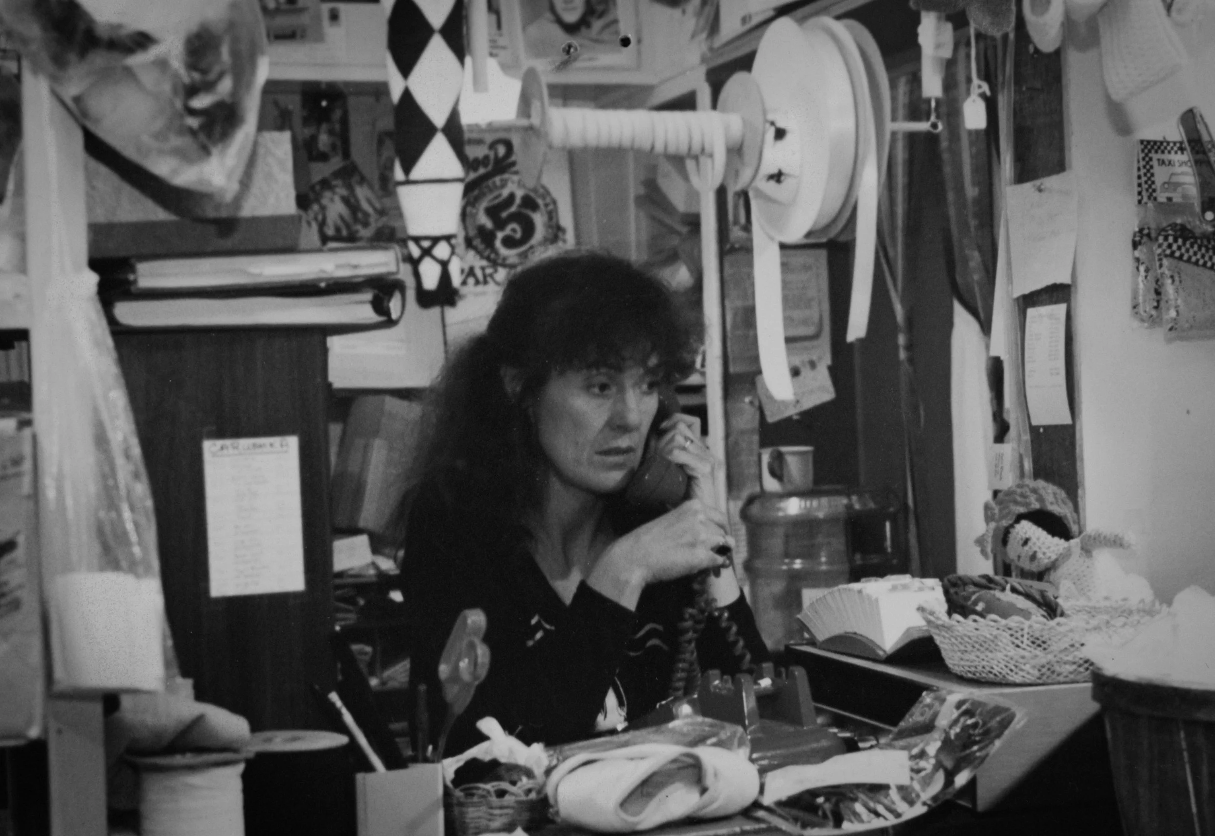 A woman with dark, wavy hair talking on a corded telephone in a cluttered office space filled with papers, a doll, books, and various office supplies.