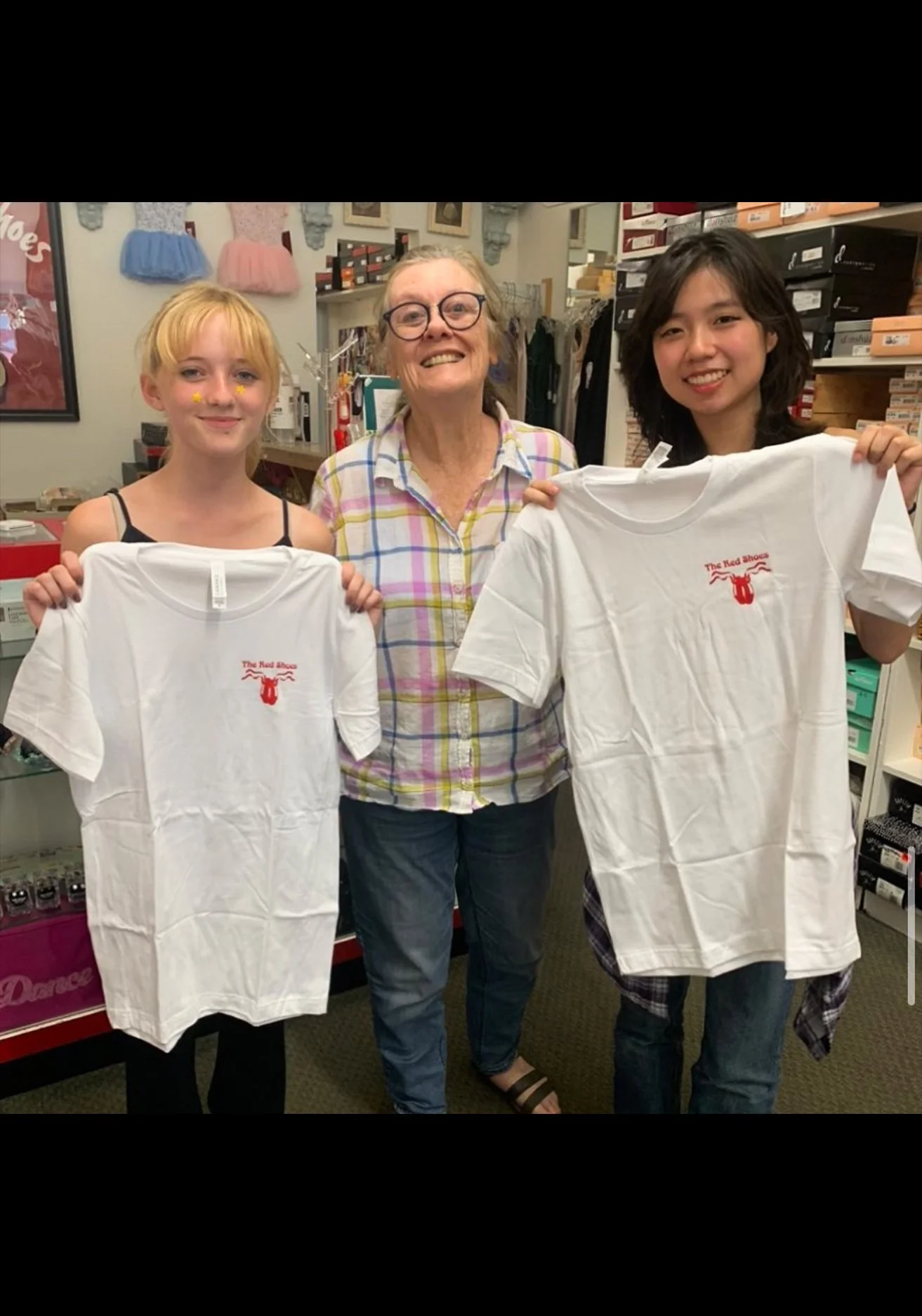 Three women standing inside a store, smiling, holding white t-shirts with red text and graphics. The women on the left and right are holding the shirts, and the woman in the middle is wearing glasses and a plaid shirt.
