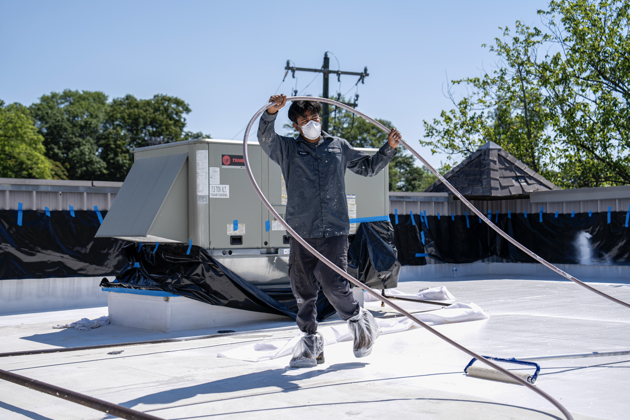 A worker wearing a face mask and protective shoe covers is painting a flat rooftop using a roller brush attached to an extension pole, with construction equipment and trees visible in the background.