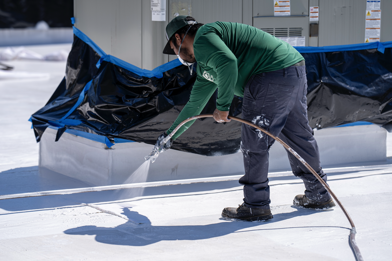 A worker wearing a green shirt, dark pants, boots, a cap, and a face mask is cleaning snow or ice from a roof with a spray hose.