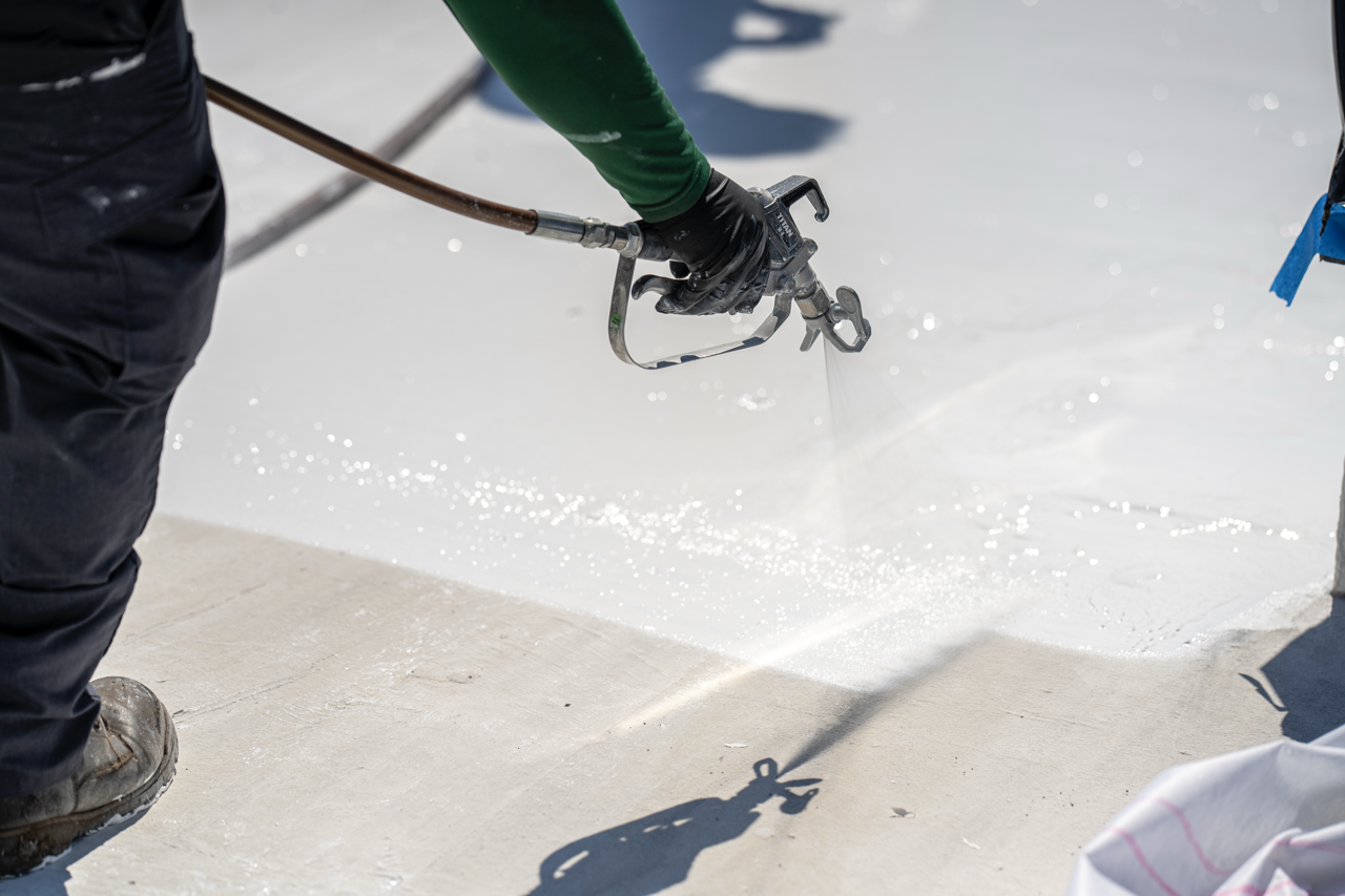 A person in black gloves and dark clothing spray painting a concrete surface white with a spray gun.