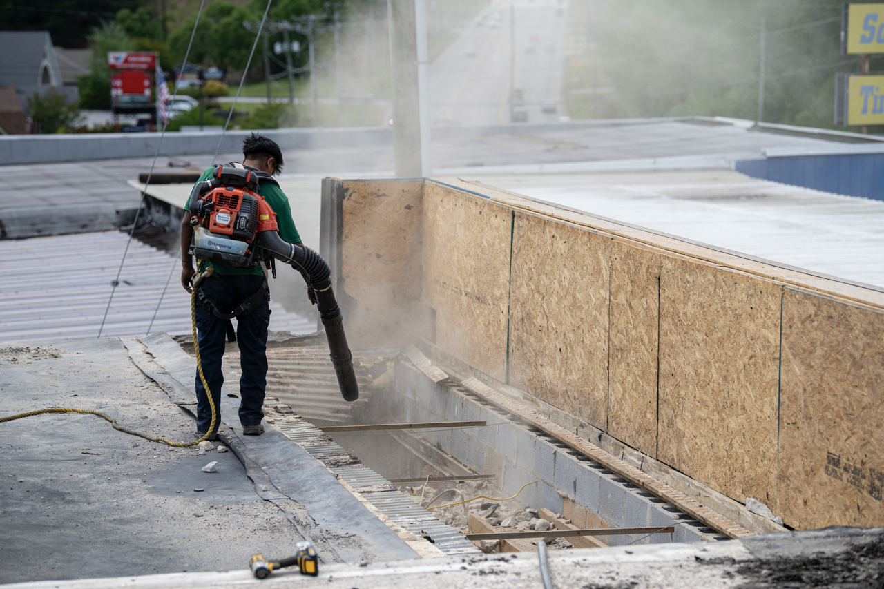 A construction worker on a rooftop using a leaf blower to clear debris. The worker wears a green shirt, black pants, and a harness, with a red and black backpack.