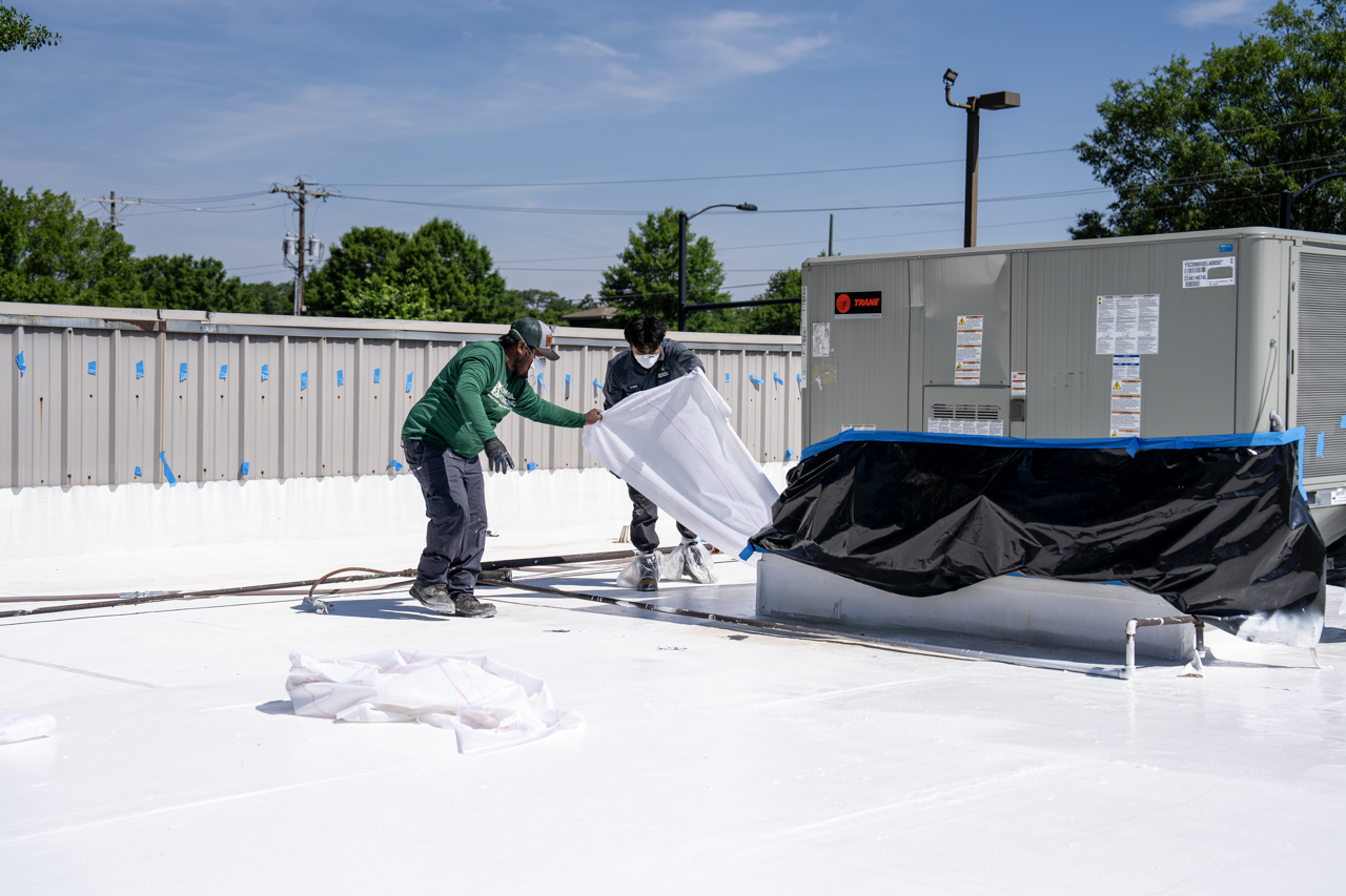 Two workers wearing masks and gloves are installing a white roofing membrane on a flat rooftop, with equipment and a fence in the background under a clear blue sky.
